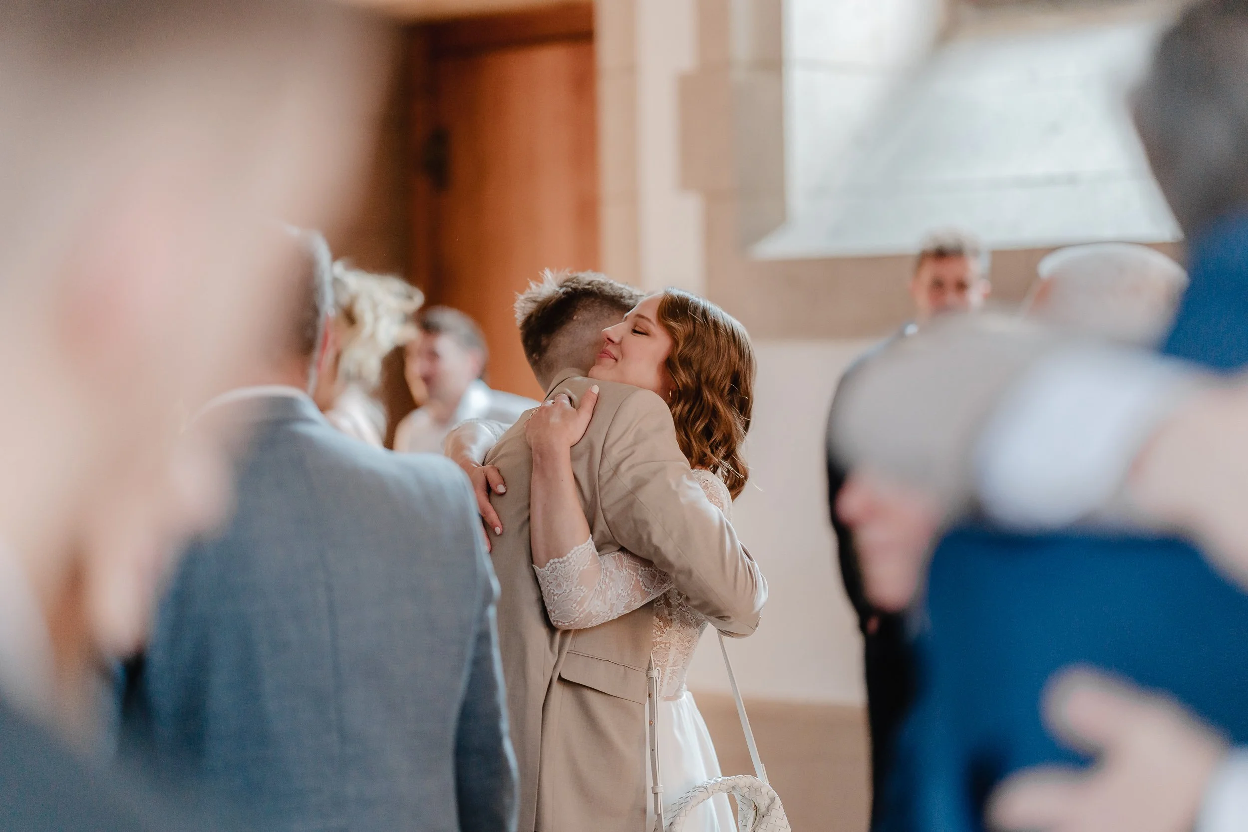 Ein Brautpaar umarmt sich liebevoll bei einer Hochzeitsfeier, umgeben von Gästen in einem festlichen Raum.