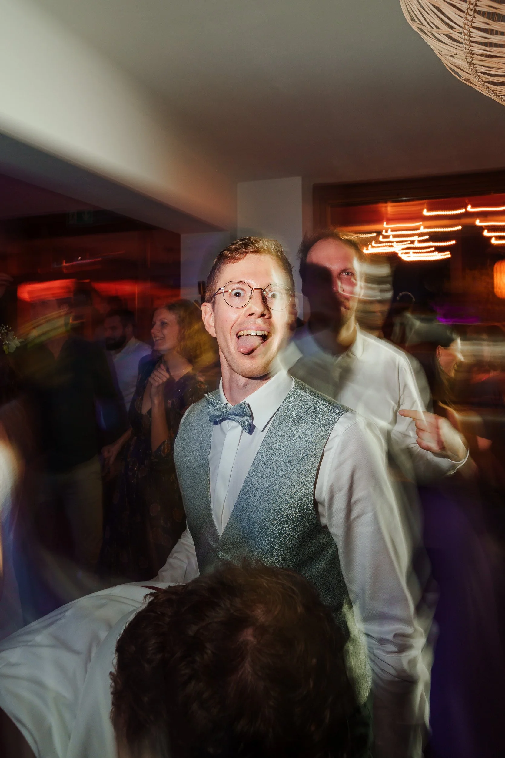 Junger Mann mit Brille und Fliege macht Grimasse auf einer Party, im Hintergrund andere Gäste, bunte Lichter und eine lebhafte Atmosphäre.