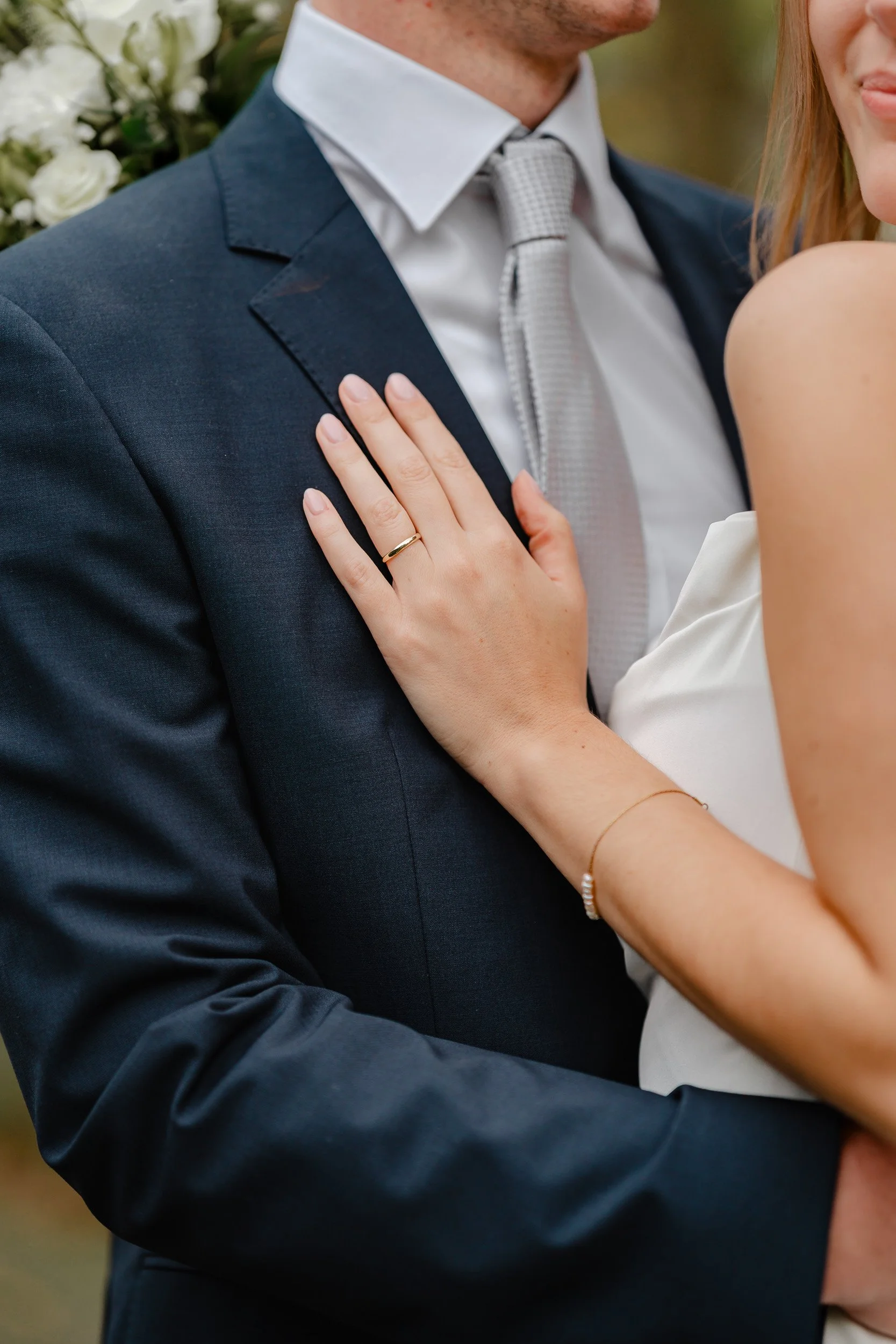 Nahaufnahme eines Paares bei einer Hochzeitszeremonie, Fokus auf die Hand der Frau mit Ehering auf dem Granatteller des Mannes, beide in formeller Kleidung, im Hintergrund Blumen.