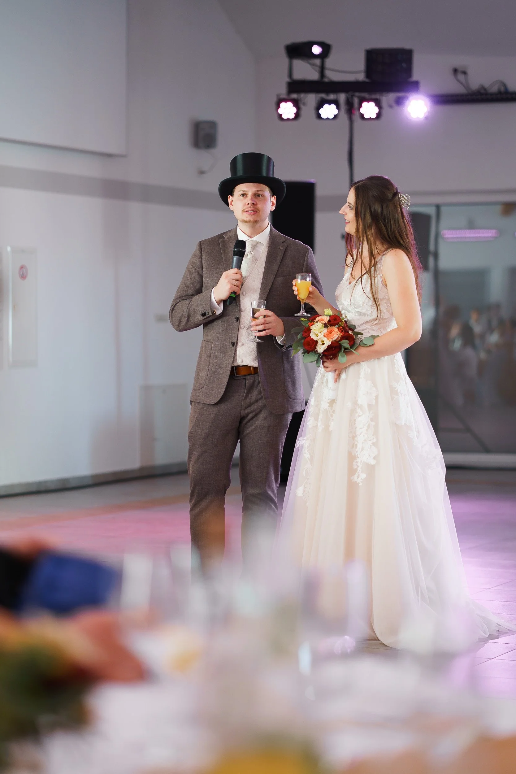 Ein Bräutigam mit Zylinderhut hält ein Mikrofon, während eine Braut mit Brautstrauß und einem Glas Orangensaft in einer Hochzeitsfeier spricht.