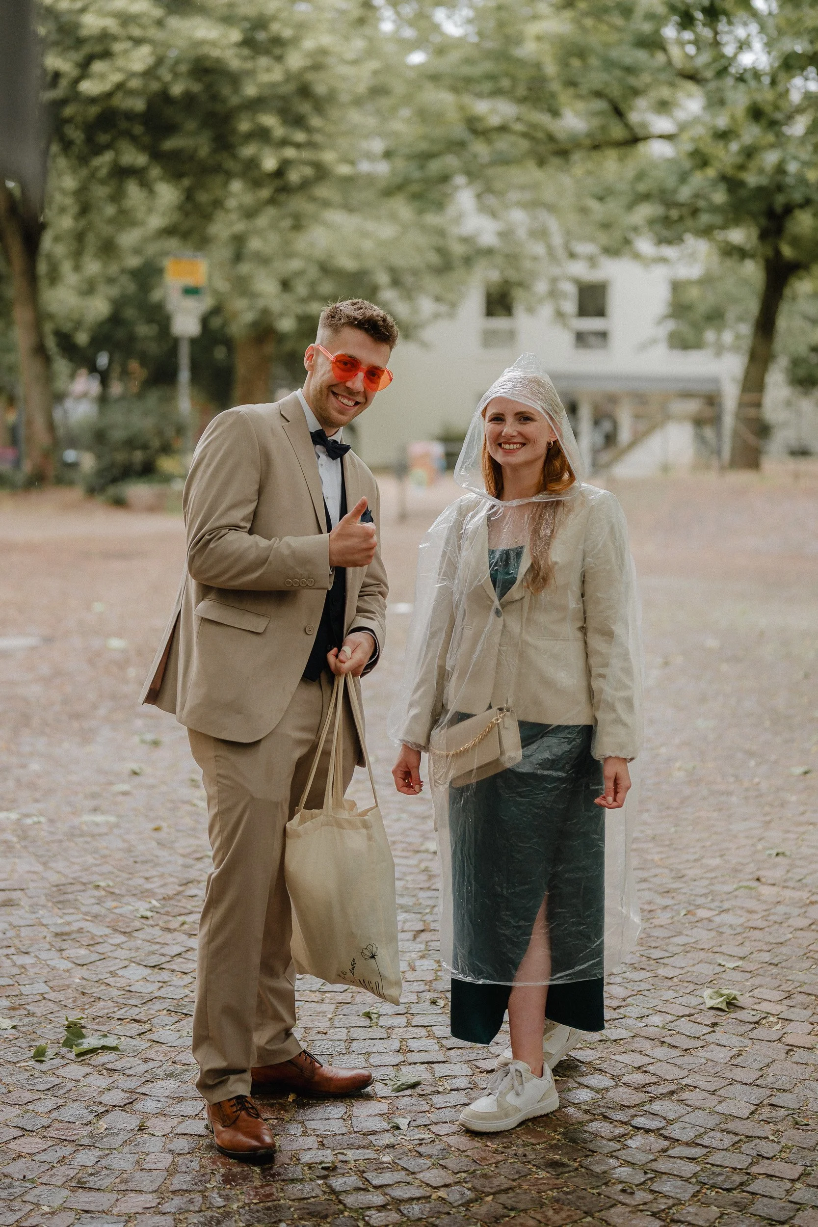 Zwei Menschen stehen auf einem gepflasterten Platz im Freien. Der Mann trägt einen beige Anzug mit weißem Hemd, schwarzem Kragen und rotem Brille und hält eine Stofftasche. Das Mädchen trägt einen schwarzen Rock, einen weißen Blazer, eine durchsichti