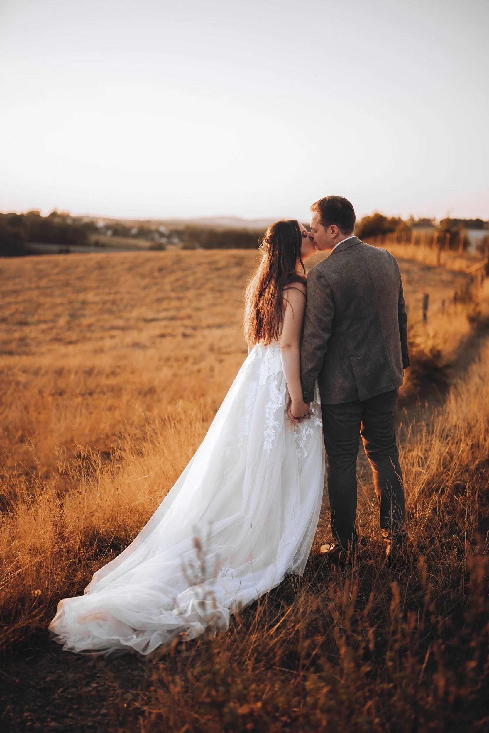 Ein Brautpaar küsst sich im Sonnenuntergang auf einem Feld, die Braut trägt ein langes weißes Kleid, der Bräutigam einen Anzug.