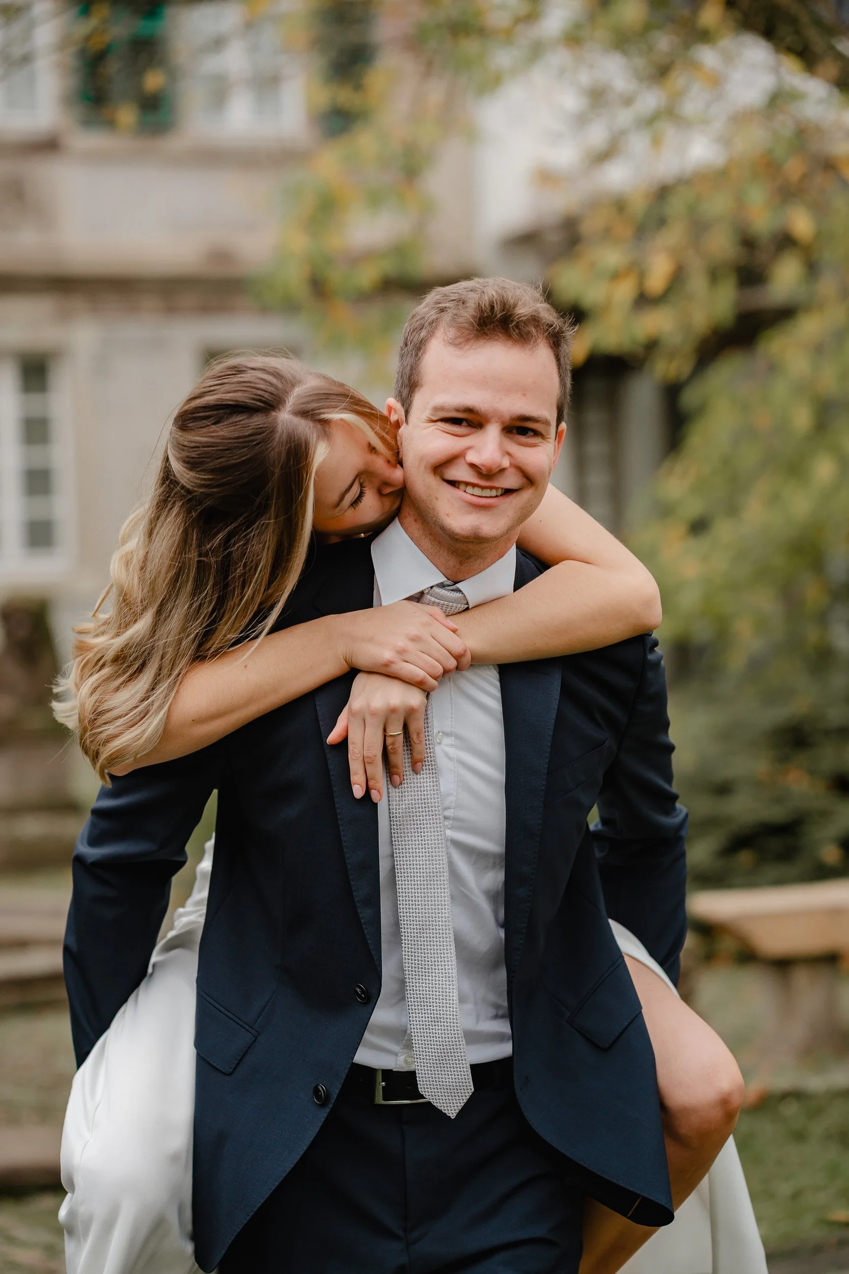 Ein lachendes Paar, der Mann trägt einen dunklen Anzug und die Frau ein weißes Kleid, im Freien mit Herbstlaub im Hintergrund.