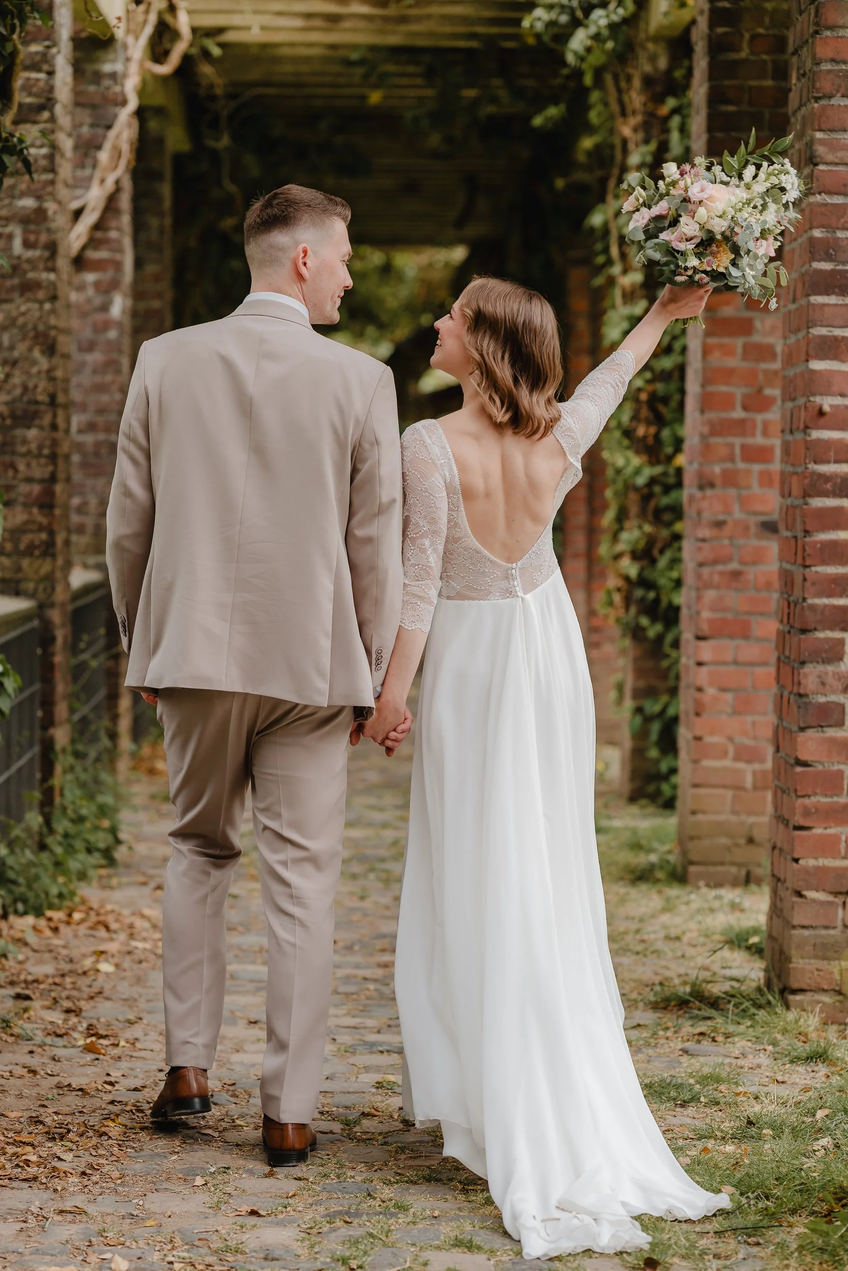 Ein Brautpaar hält Händchen, sie trägt ein weißes Hochzeitskleid mit Spitze und hebt einen Blumenstrauß. Sie gehen auf einem gepflasterten Weg zwischen roten Ziegelwänden und grünen Pflanzen.