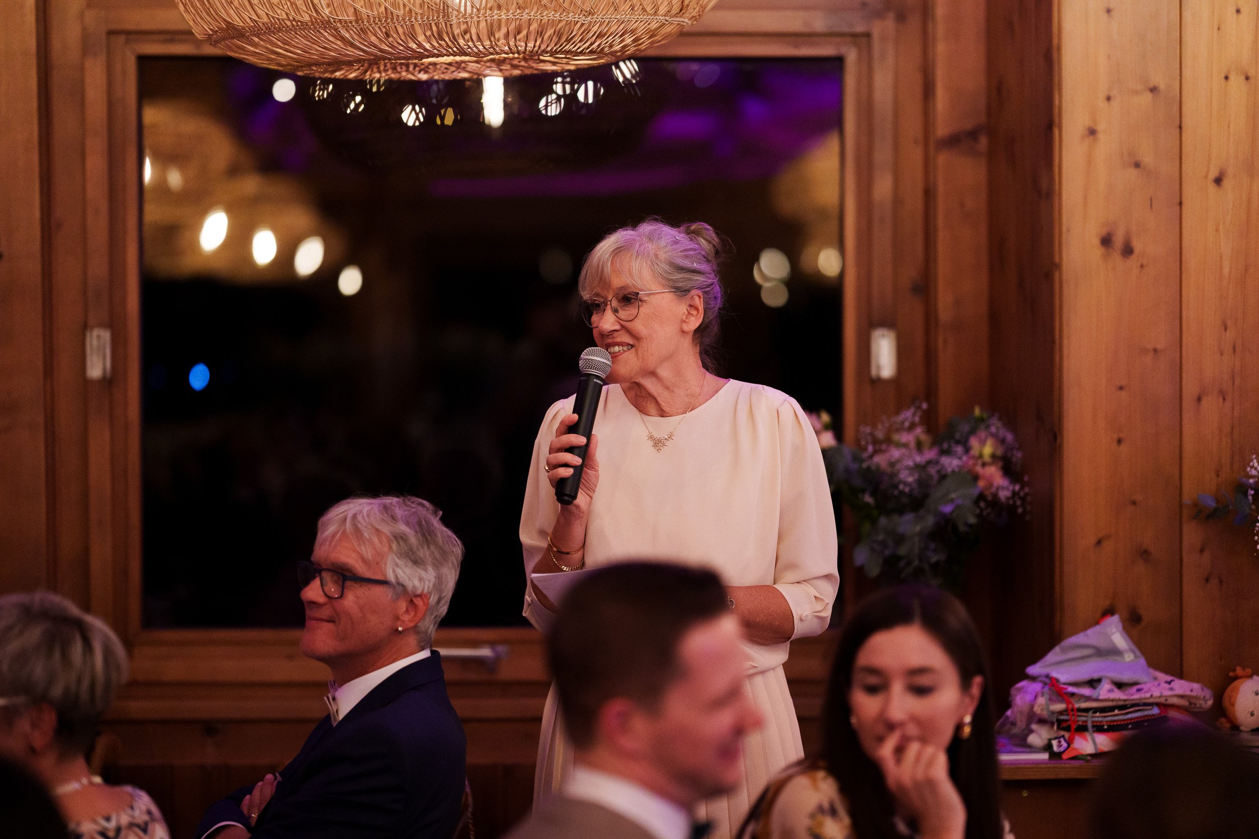 Eine ältere Frau mit grauen Haaren und Brille hält eine Mikrofon beim Reden bei einer Feier. Sie trägt ein cremefarbenes Kleid und steht vor einer Gruppe von Menschen in einem rustikalen Holzraum mit Blumen und Lampen.