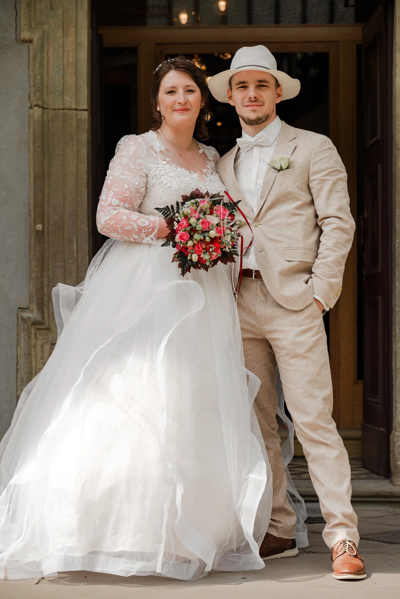 Braut und Bräutigam stehen vor einer Tür, die Braut hält einen Blumenstrauß, der Bräutigam trägt einen weißen Hut und einen beige Anzug.