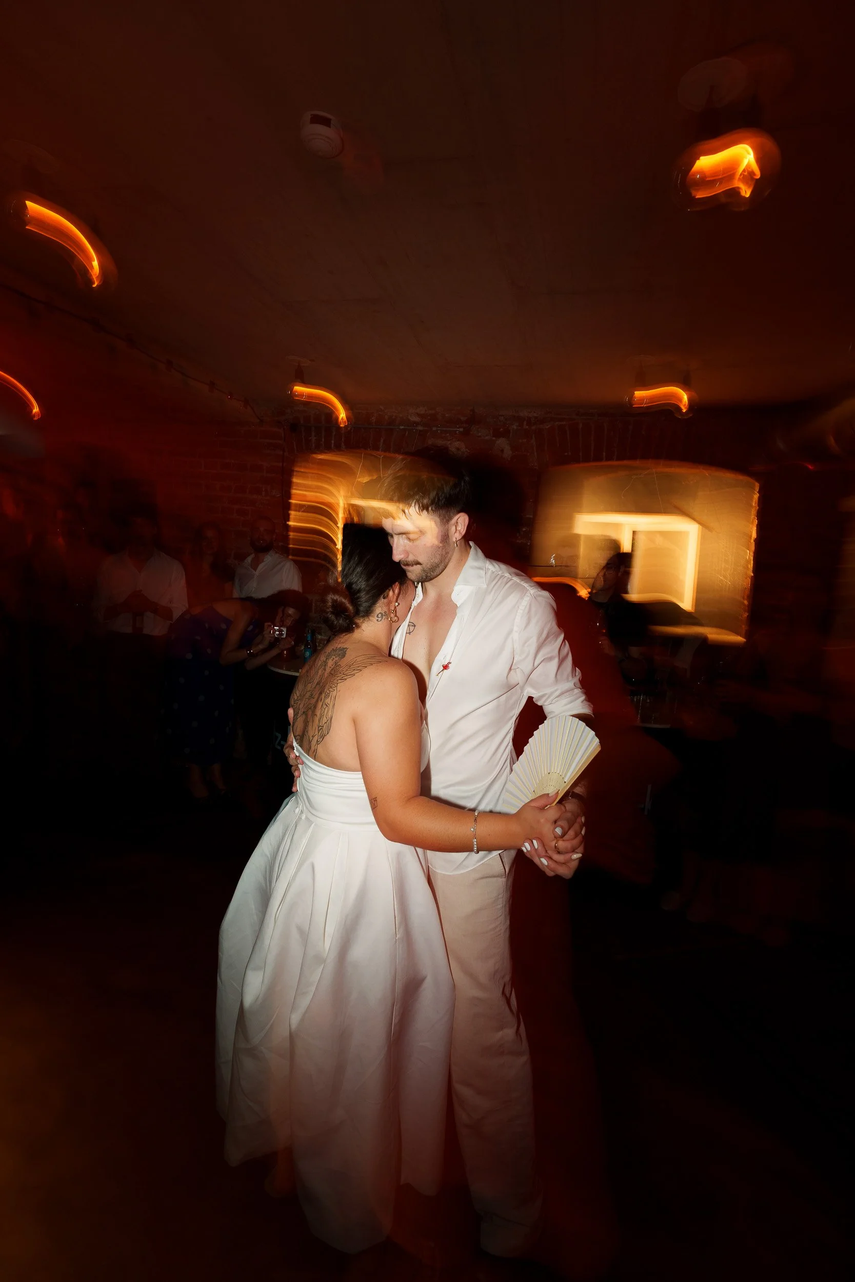 Ein Paar tanzt eng umgeben von lauter Menschen in einem dunklen Raum mit warmem Licht und Bewegungsunschärfe.