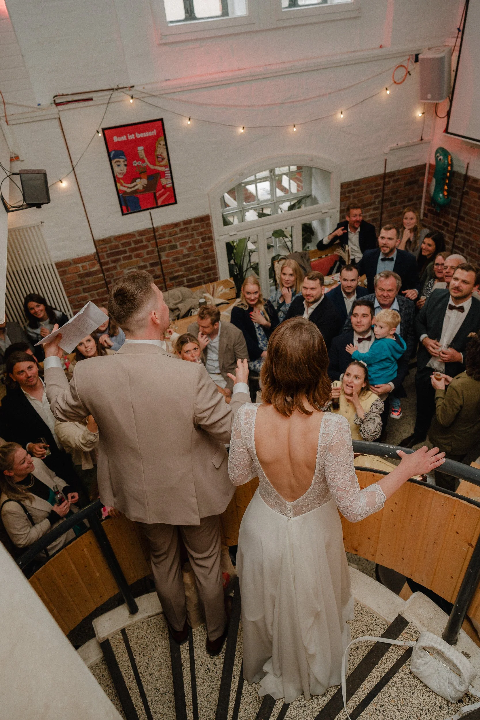 Ein Hochzeitspaar steht auf einer Treppe, vor einem Publikum in einem festlich dekorierten Raum, während die Braut ein langärmeliges, off-shoulder Hochzeitskleid trägt. Das Publikum schaut ihnen zu, während der Bräutigam eine Rede hält.
