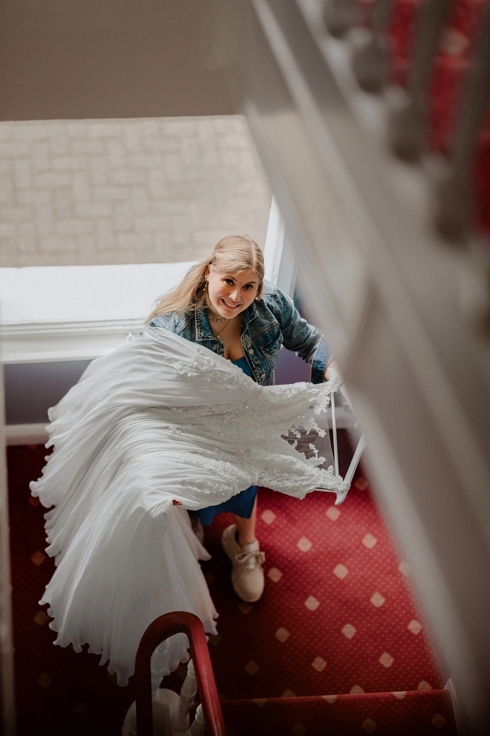 Getting Ready | Gummersbach | Junge Frau schaut beim Aufstieg die Treppe hinunter, hält ein Hochzeitskleid in der Hand, trägt eine Jeansjacke, blaue Kleidung darunter und Turnschuhe, im Haus.