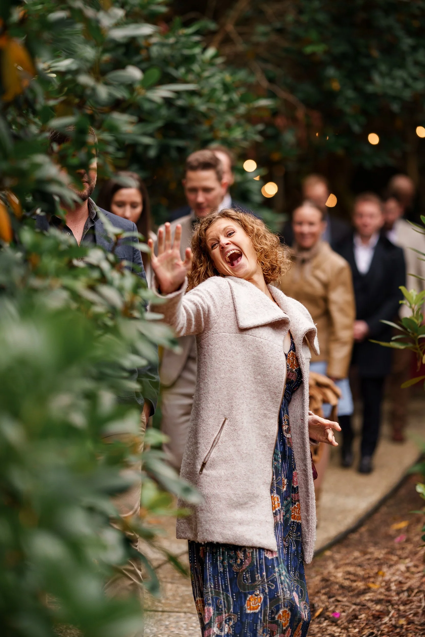 Fröhliche Frau winkt und lacht, während sie durch eine Hecke auf einem Gartenweg geht, umgeben von anderen lachenden und fröhlichen Menschen.