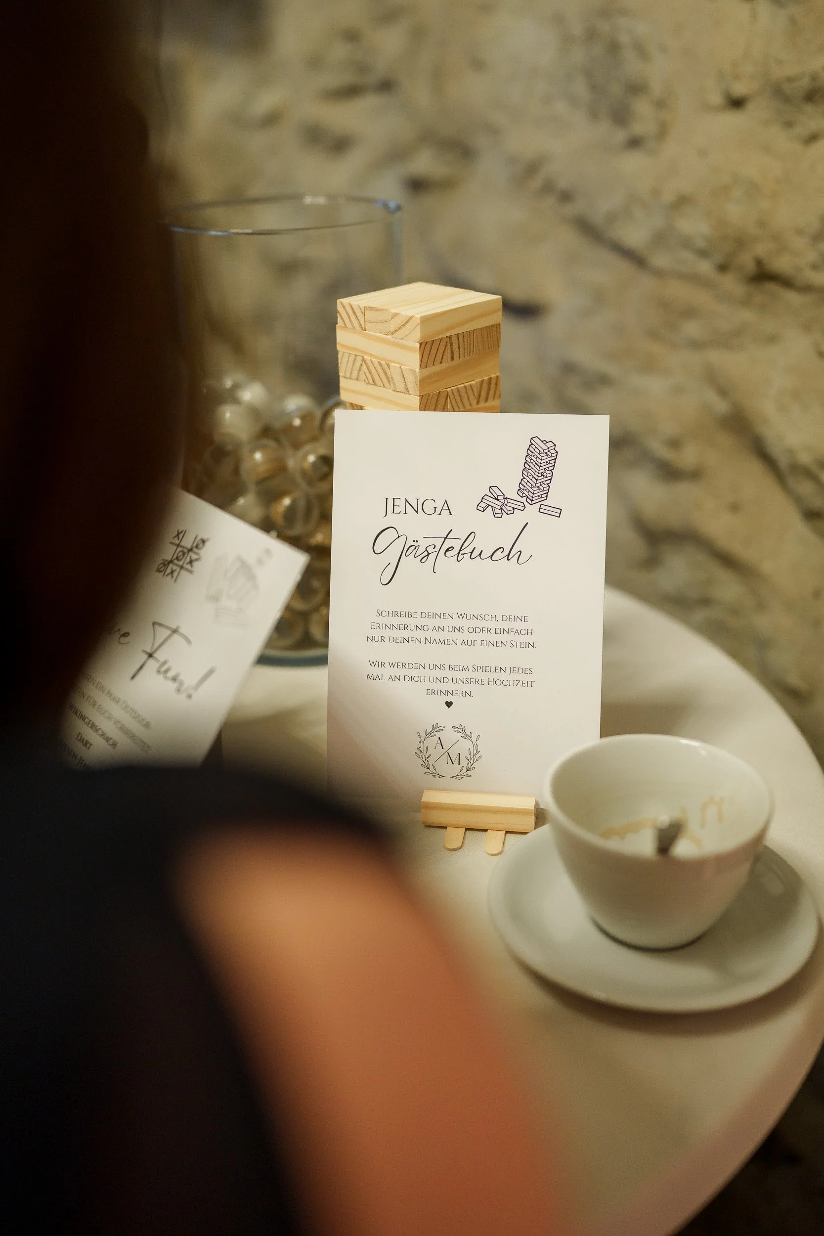 Ein Gästebucheintrag für Jenga-Spiele bei einer Feier, mit einer kleinen Holzpyramide und einer Tasse auf einem Tisch, im Hintergrund eine Glasvase mit Steinen, vor einer Steinmauer.