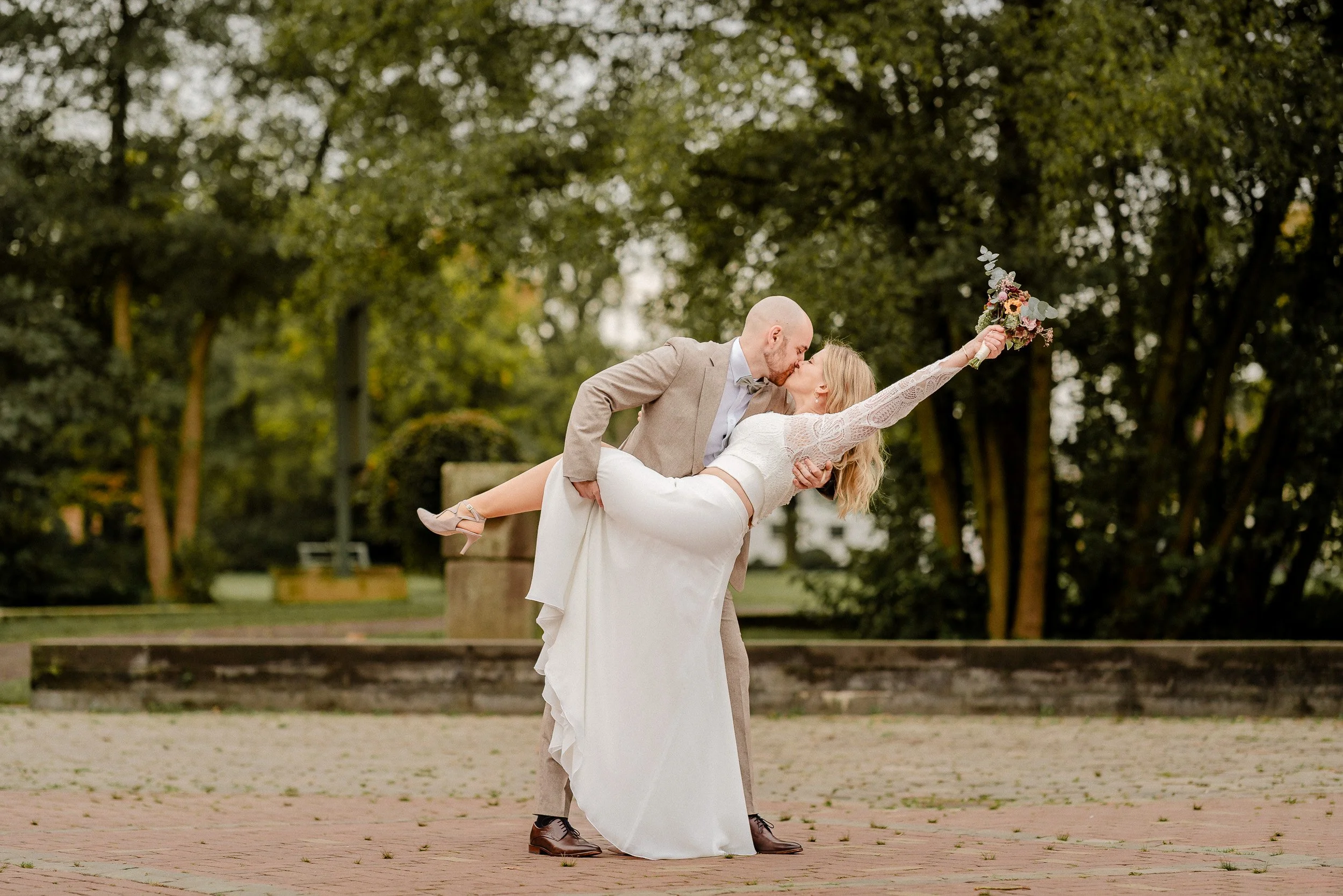 Ein Brautpaar bei einer Hochzeit im Freien, der Bräutigam hält die Braut in den Armen, sie küsst sich, die Braut hält einen Blumenstrauß in die Luft.
