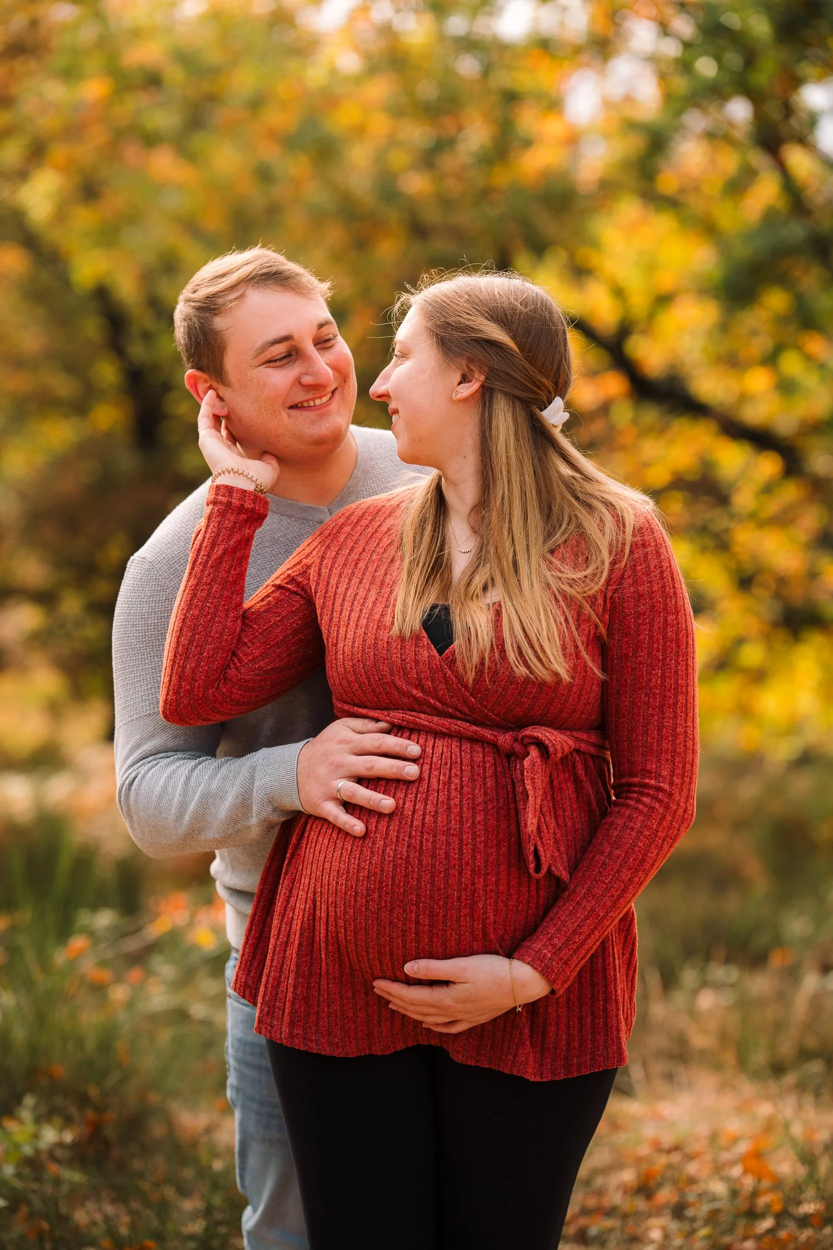 Ein Paar steht im Park im Herbst, die schwangere Frau trägt eine rot-orange Strickjacke, der Mann hält sie und küsst sie zärtlich, beide lachen und schauen sich liebevoll an.