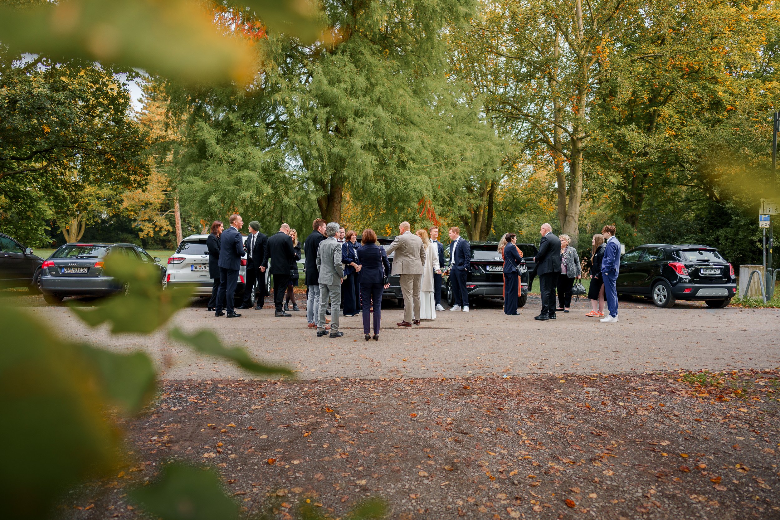Gruppen von Menschen bei einer Feier im Freien, umgeben von Bäumen, mit mehreren Autos im Hintergrund.