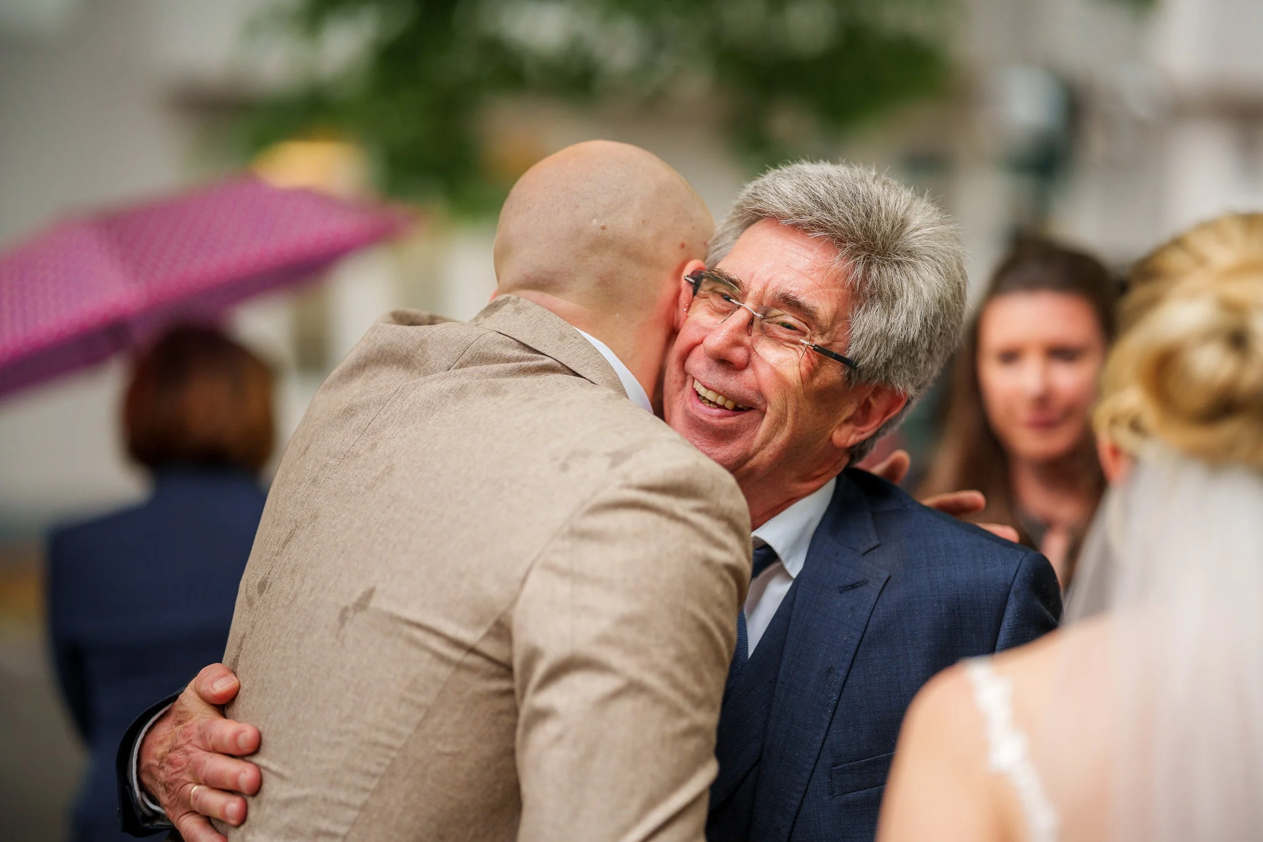 Zwei Männer umarmen sich beim Empfang, lächelnd, mit anderen Menschen im Hintergrund, bei schlechtem Wetter mit einem pinken Regenschirm.