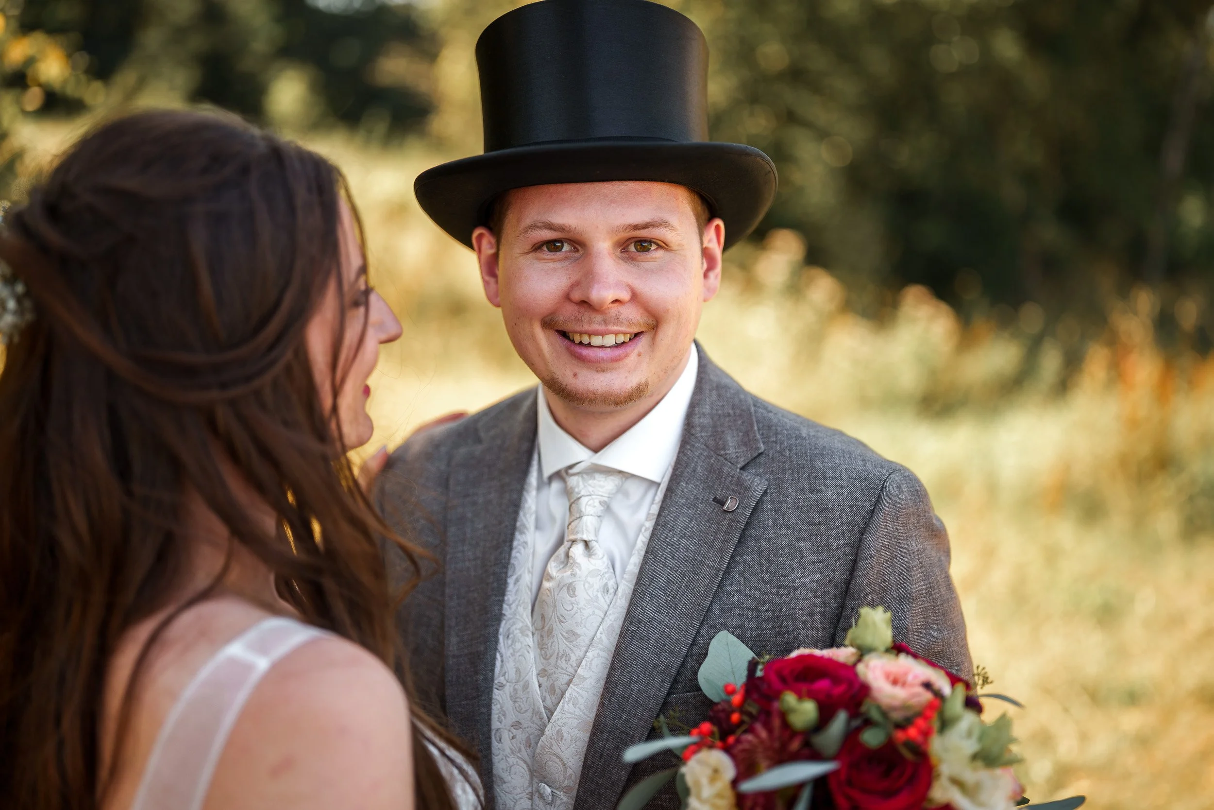 Ein glückliches Brautpaar bei einer Hochzeit im Freien. Der Bräutigam trägt einen schwarzen Zylinder und einen grauen Anzug, während die Braut einen weißen Brautkleid trägt und einen Blumenstrauß hält.