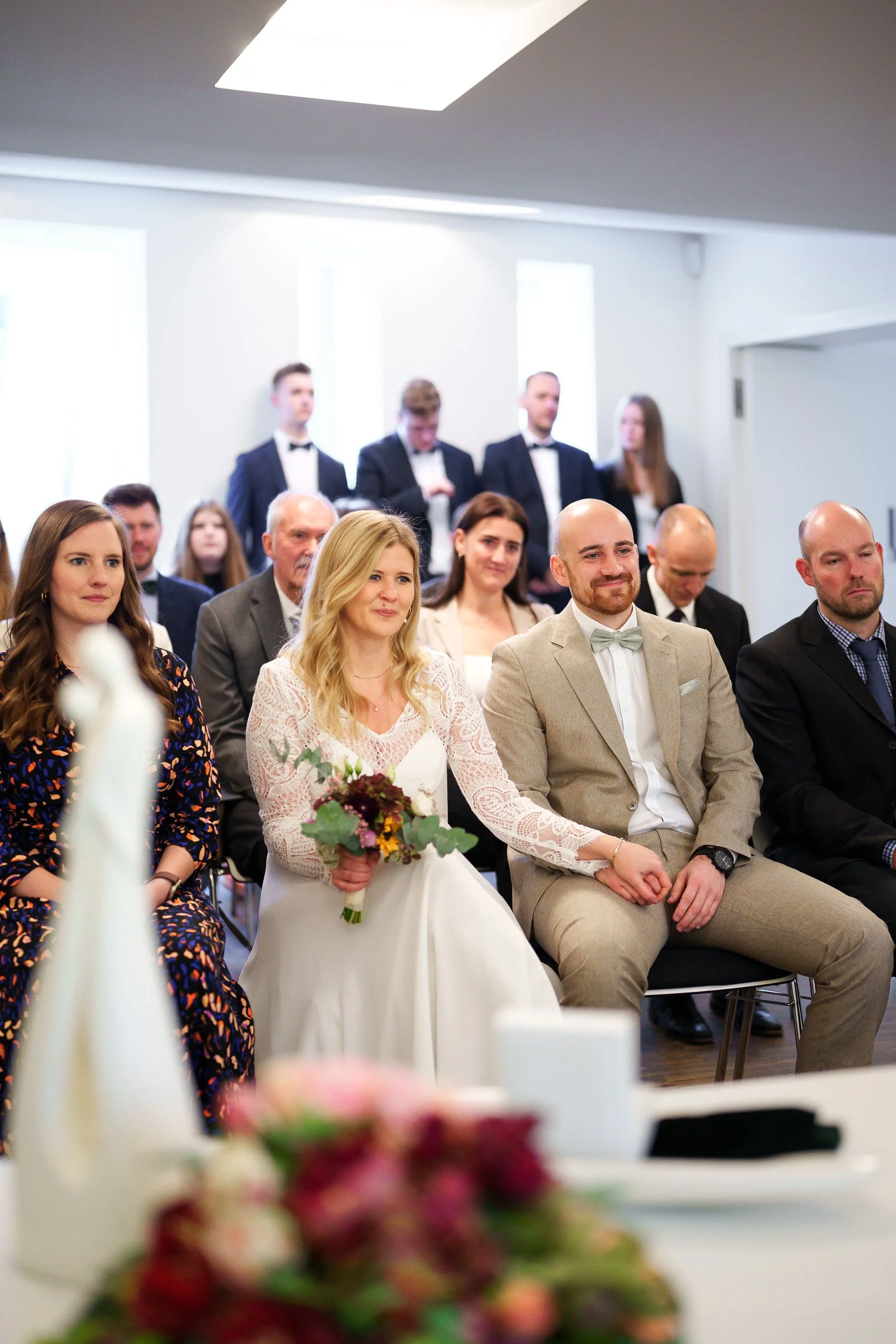 Eine Gruppe von Menschen bei einer Hochzeitszeremonie, sitzend in einem festlichen Raum, mit Braut in weißem Kleid und Bräutigam in beige Anzug.