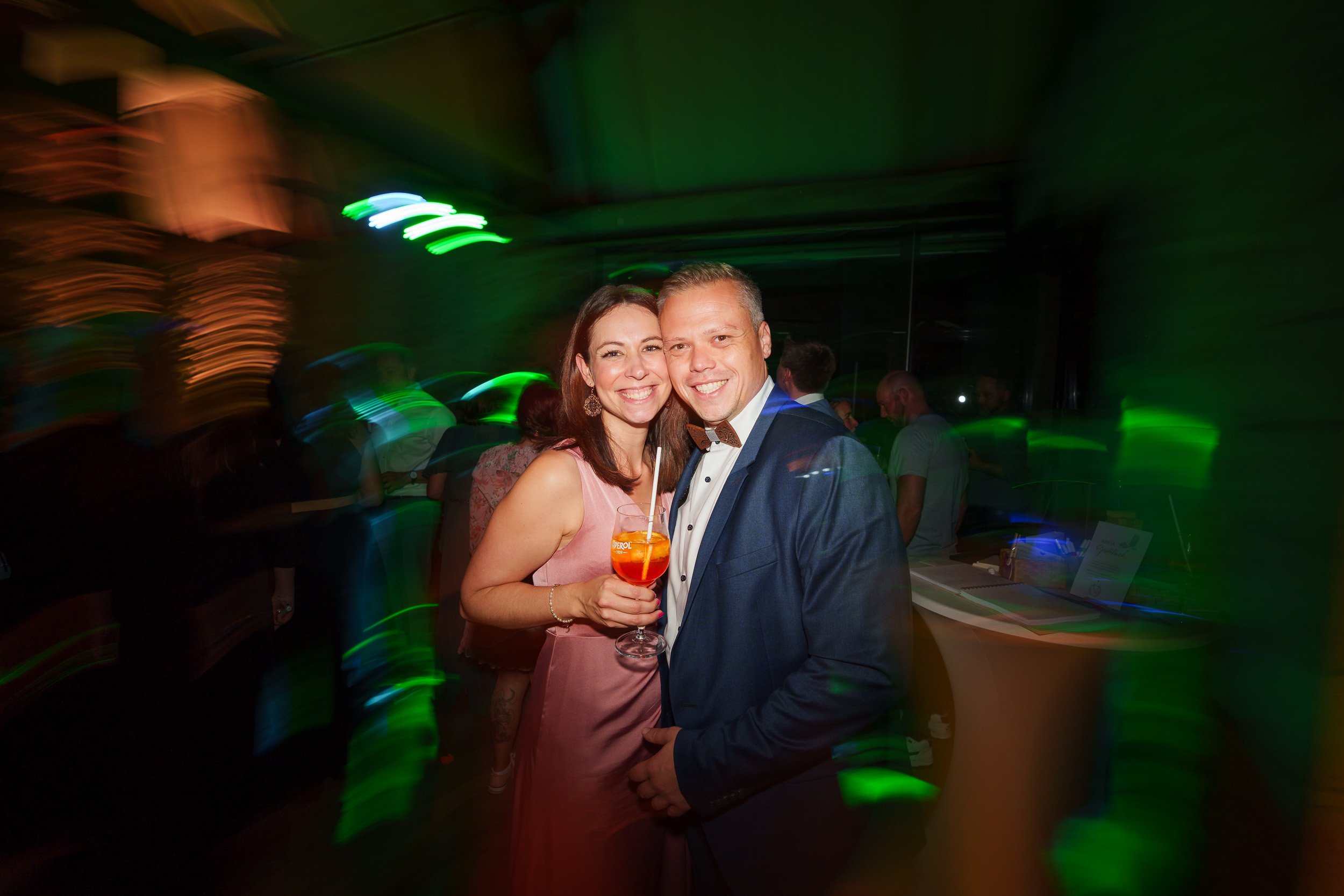 Ein lachendes Paar in festlicher Kleidung bei einer Party, der Frau hält ein Getränk in der Hand, im Hintergrund sind tanzende Menschen und bunte Lichter.