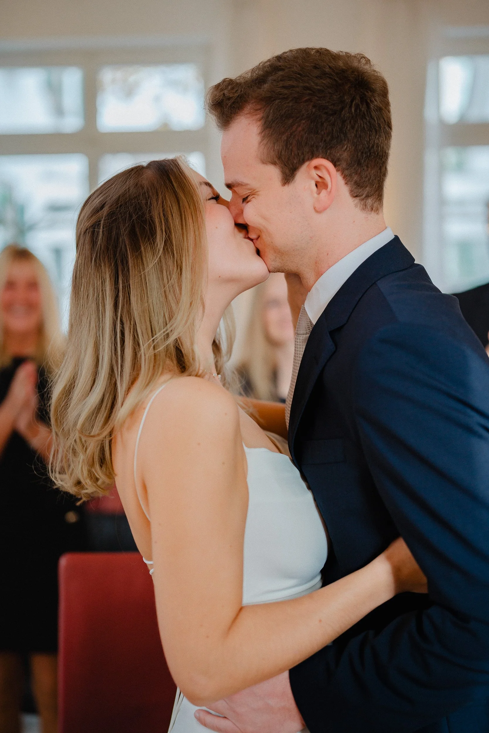 Ein Mann und eine Frau küssen sich bei einer Hochzeit, im Hintergrund applaudierende Gäste, helle Fenster im Raum.