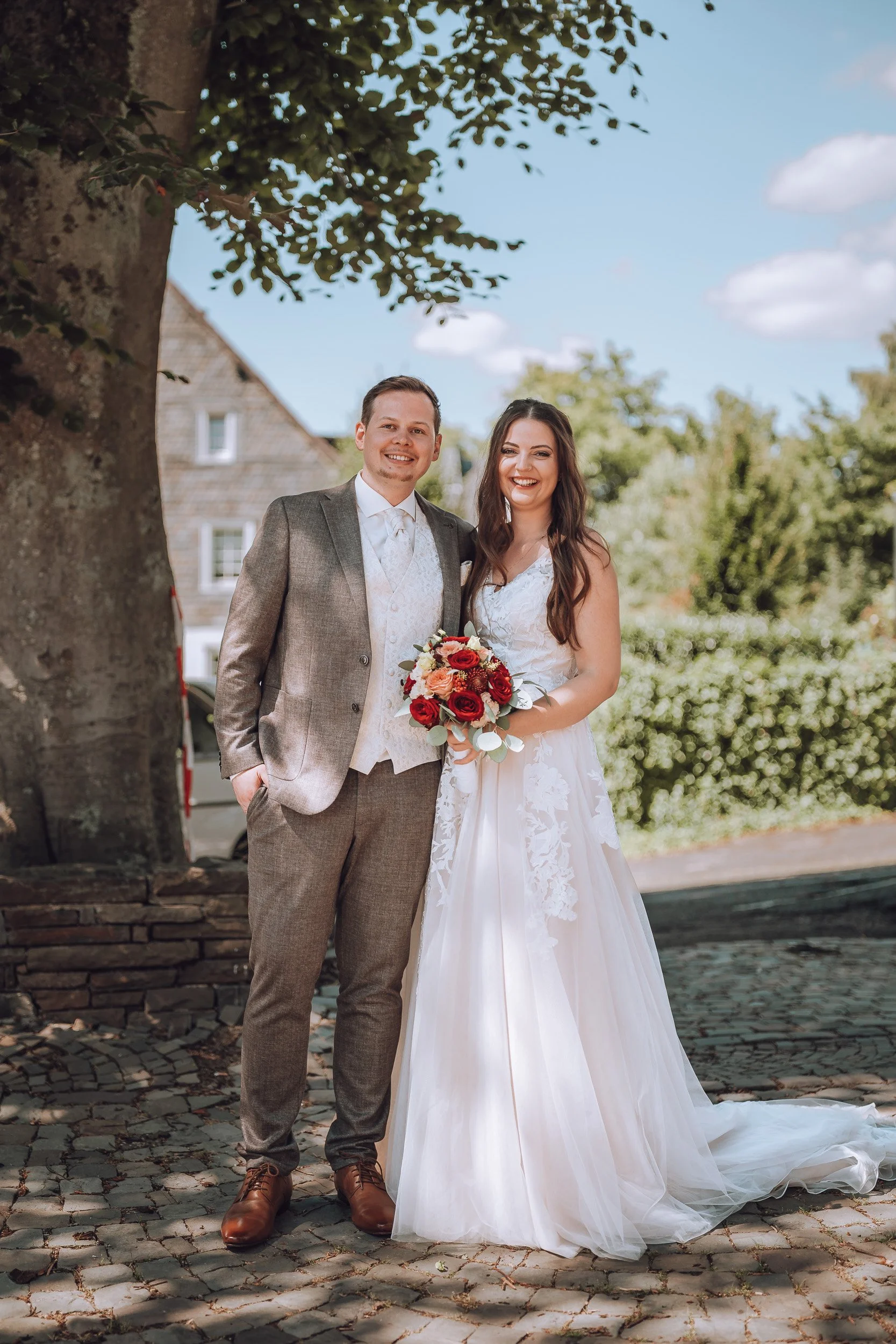 Ein frisch verheiratetes Paar steht lachend vor einem Baum, die Braut hält einen Blumenstrauß mit roten und weißen Blumen, draußen bei schönem Wetter.