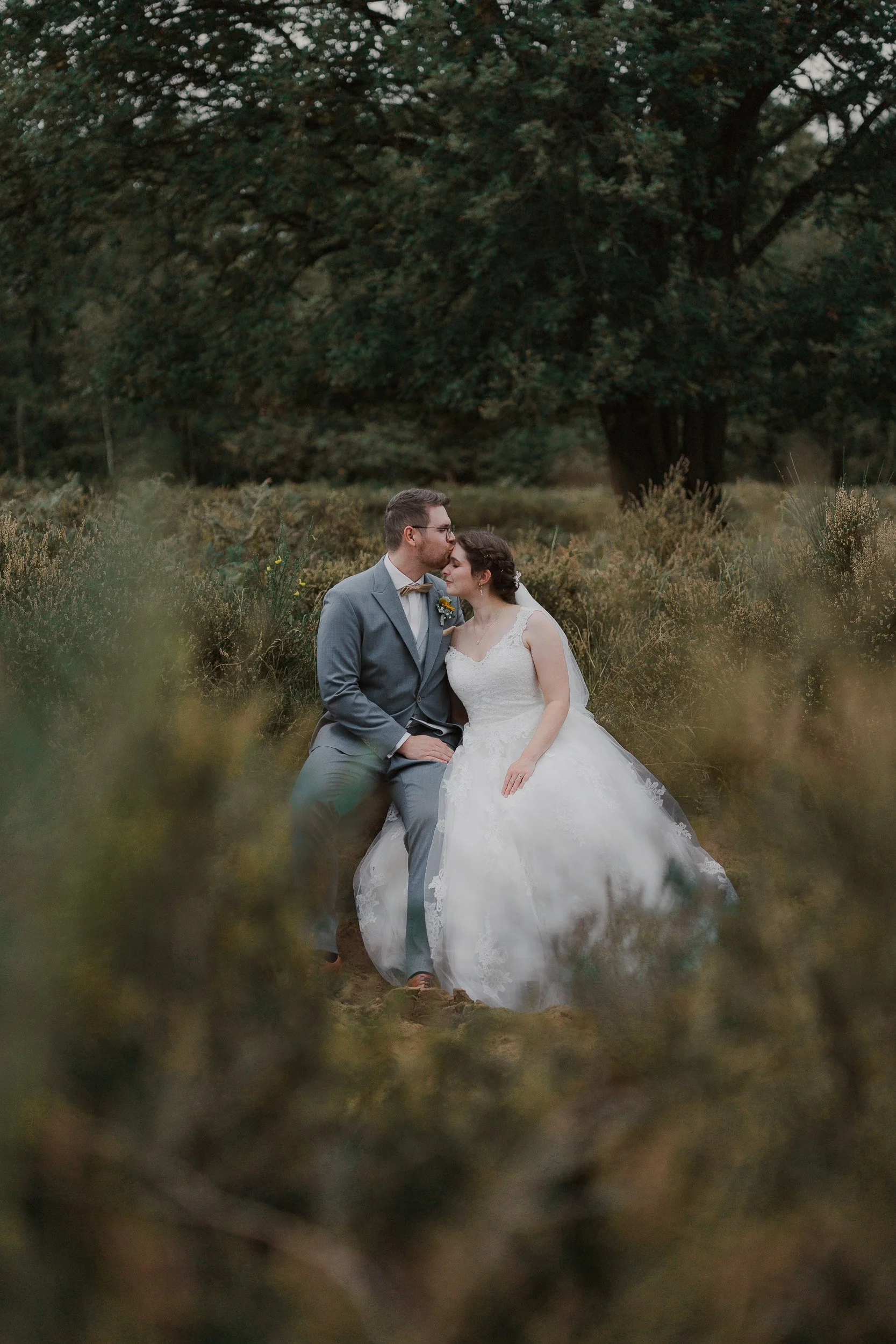Ein Brautpaar sitzt im Grünen, der Bräutigam küsst die Braut auf die Stirn, im Wald umgeben von Büschen und Bäumen.