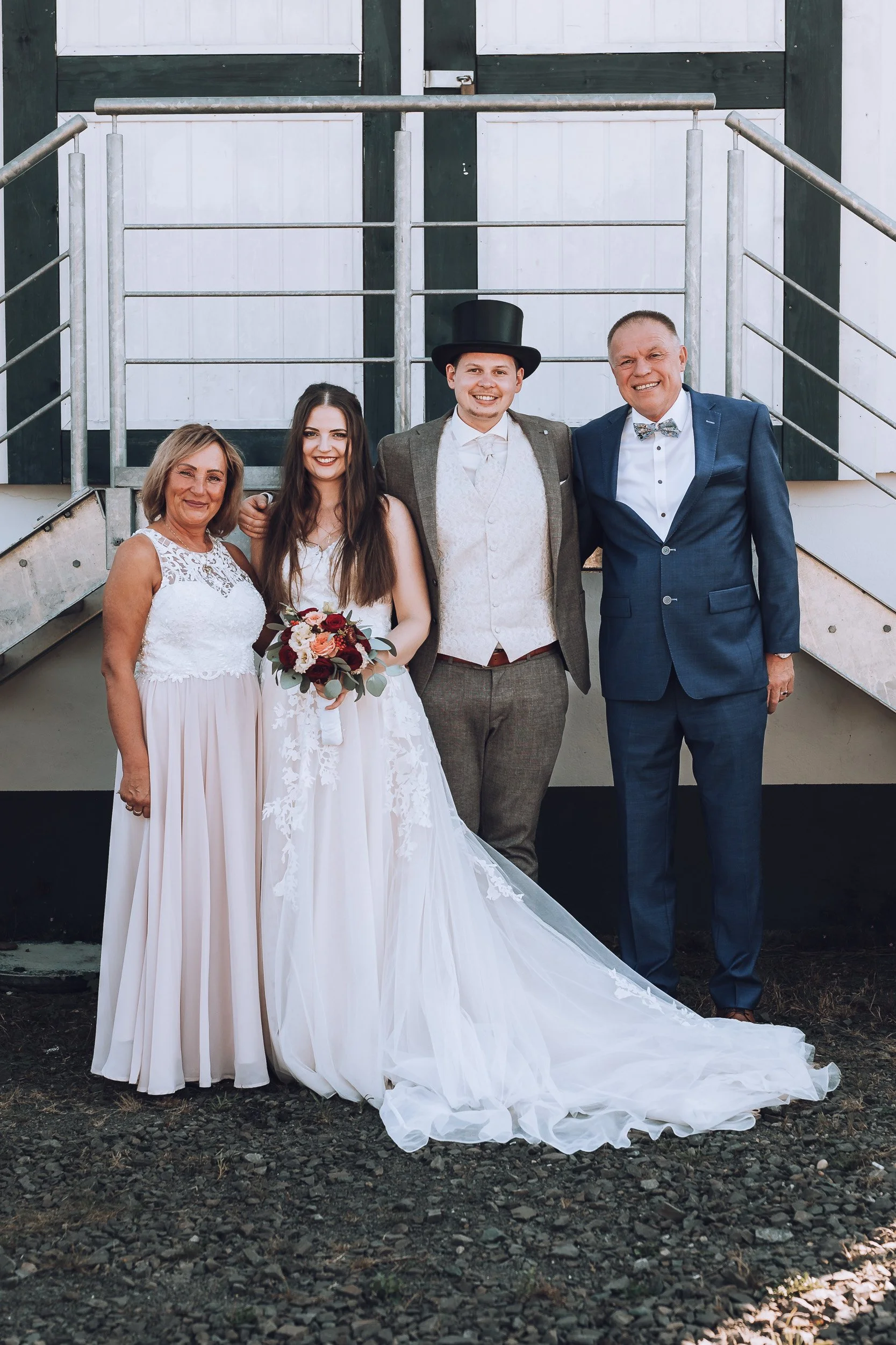 Vier Personen auf einer Hochzeit, eine Braut in einem weißen Brautkleid mit Blumenstrauß, zwei Männer in Anzügen, und eine ältere Frau in einem weißen Kleid, vor einer Industrie- oder Scheunentrennwand.