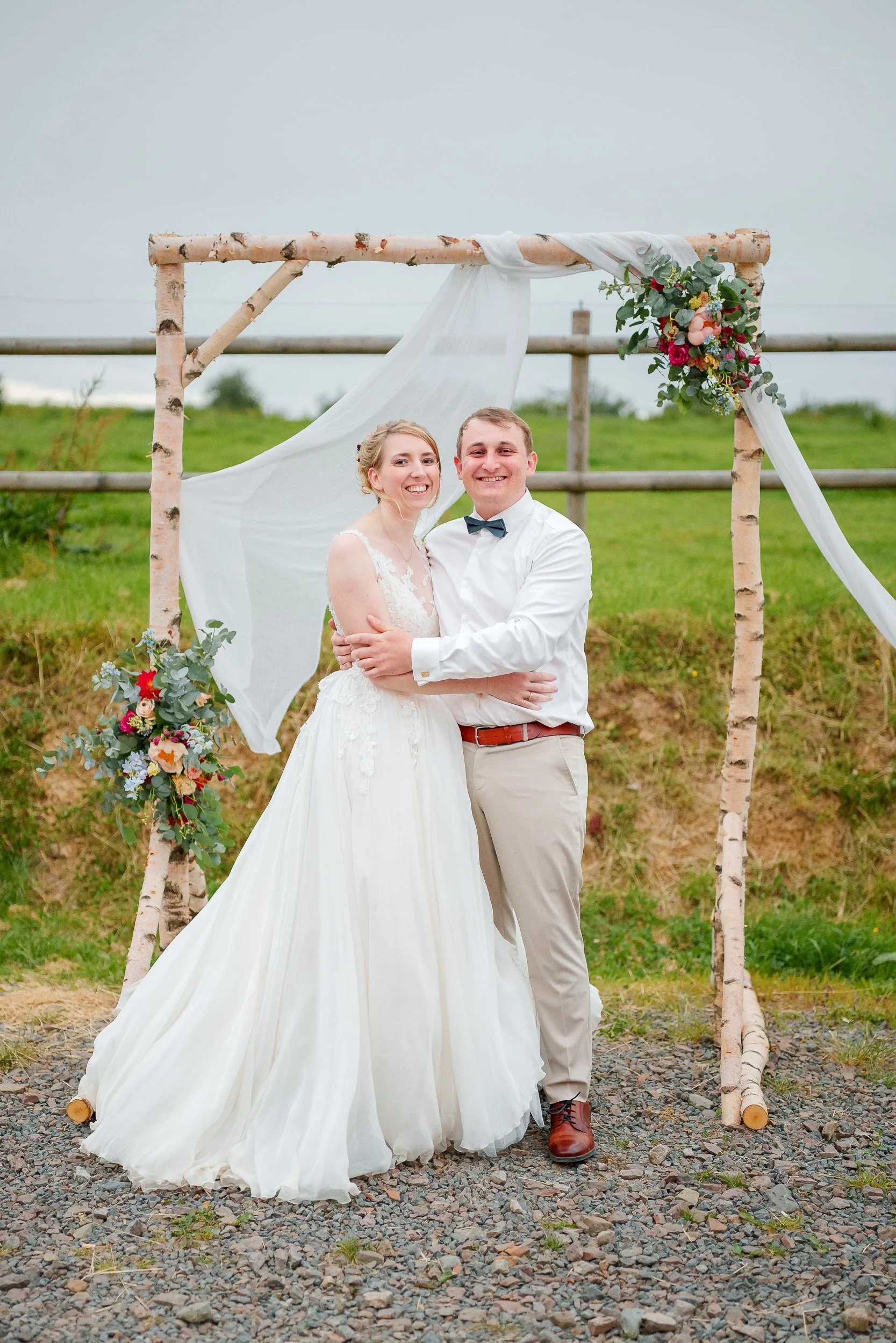 Ein glückliches Brautpaar im Hochzeitsoutfit, umarmt sich vor einem holzgestützten Altar mit weißen Stoffen und Blumen, im Freien auf einer Wiese bei bewölktem Himmel.