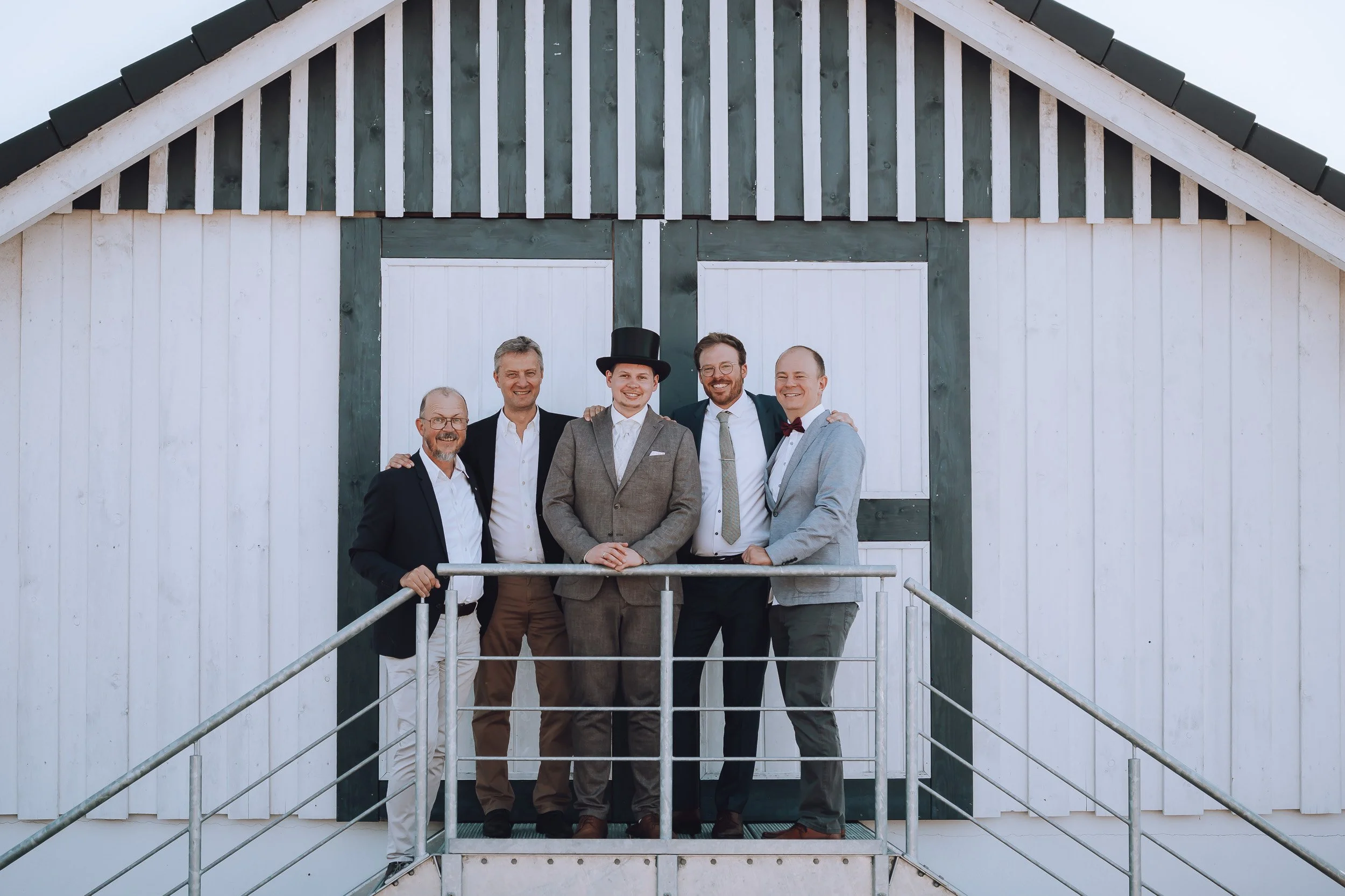 Fünf Männer in formeller Kleidung stehen auf einer Metallplattform vor einer weißen Scheune mit grünen Akzenten, einige lachen und haben Arme um die Schultern der anderen.