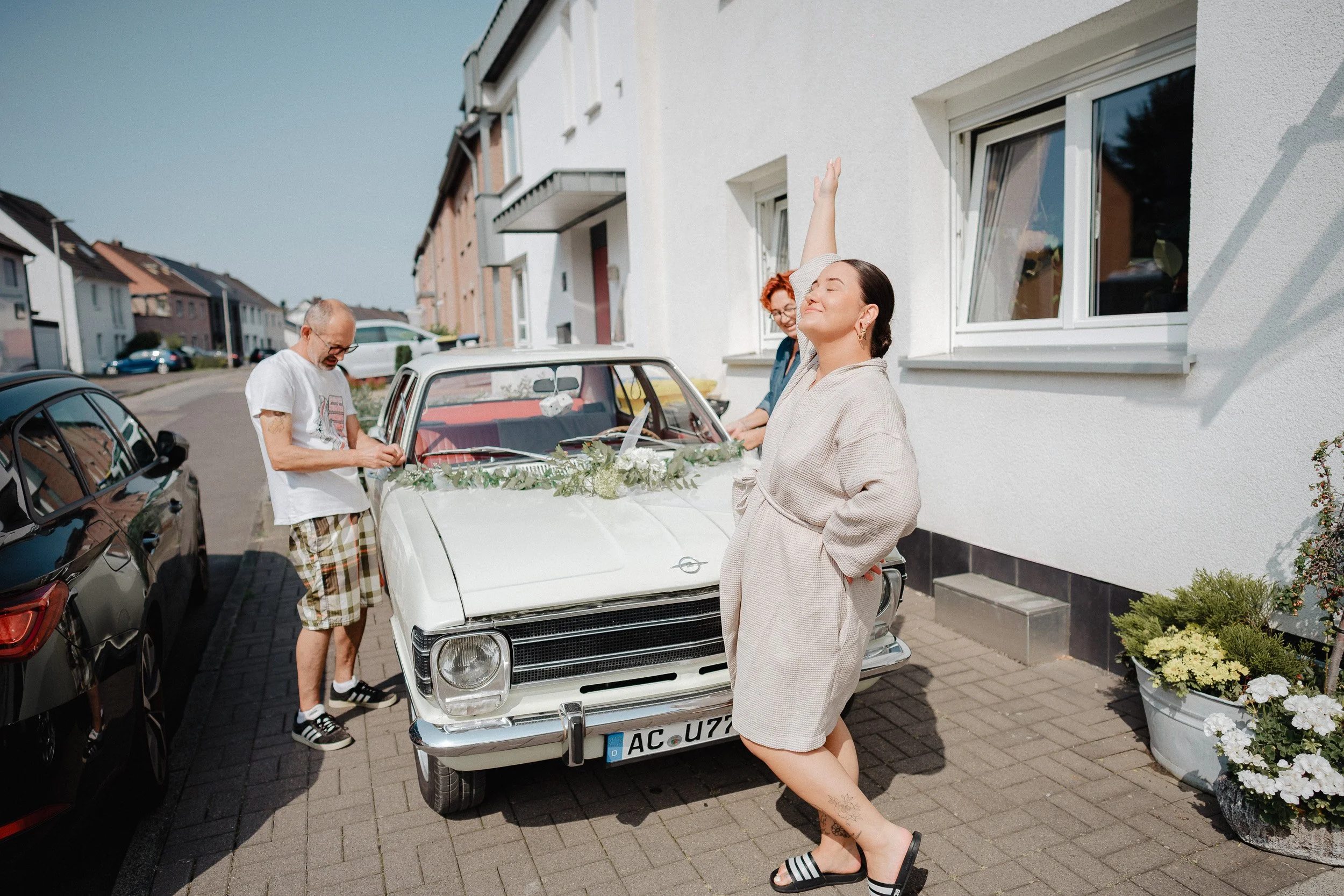 Drei Personen feiern neben einem Oldtimer, der mit Blumen dekoriert ist, vor einem weißen Haus auf einem Bürgersteig.