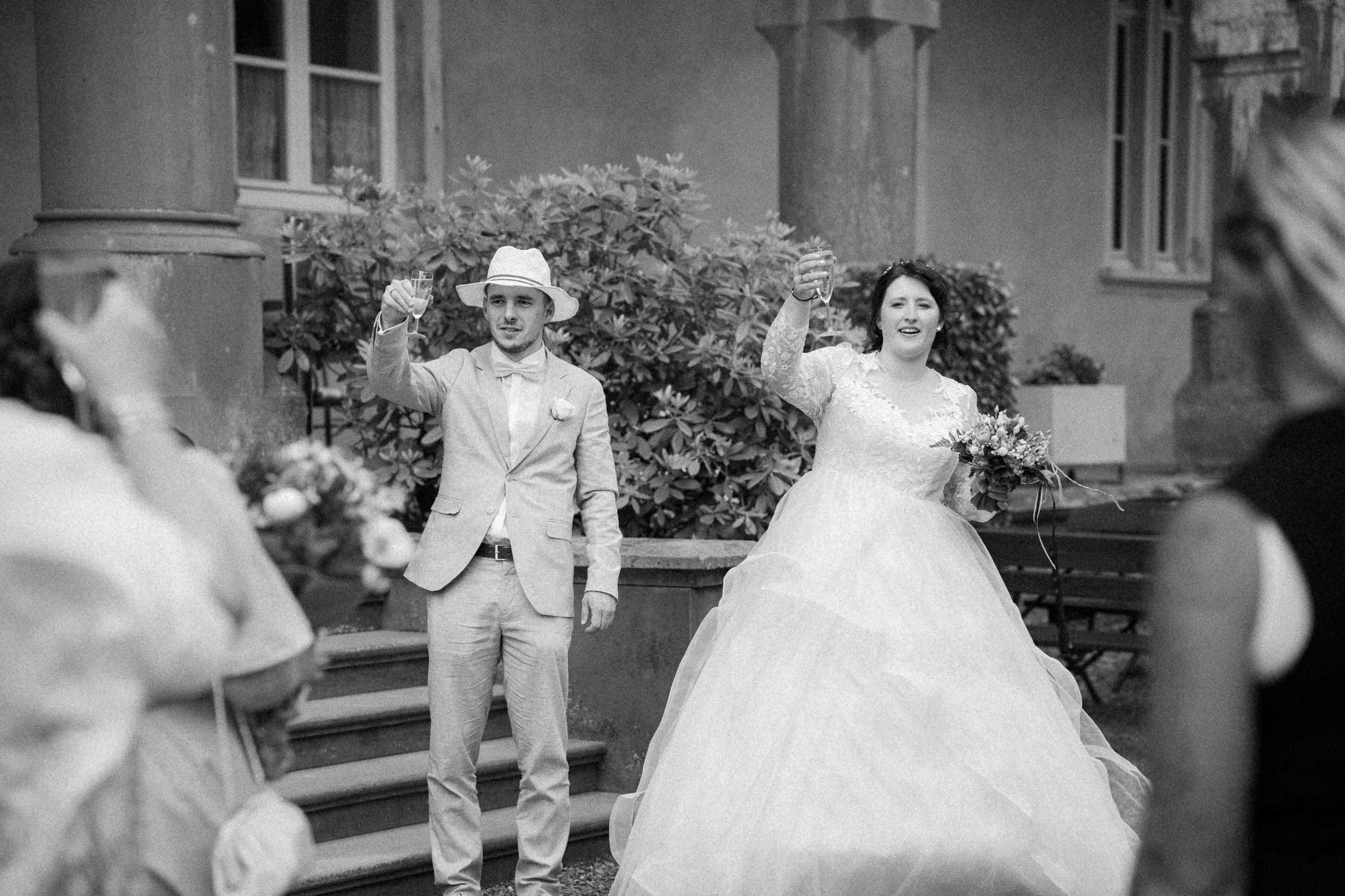 Ein Brautpaar bei einer Hochzeit, Getönte schwarze und weiße Aufnahme, hebt Gläser zum Anstoßen, umgeben von Gästen, im Außenbereich vor einem Gebäude mit Fenstern und Pflanzen.