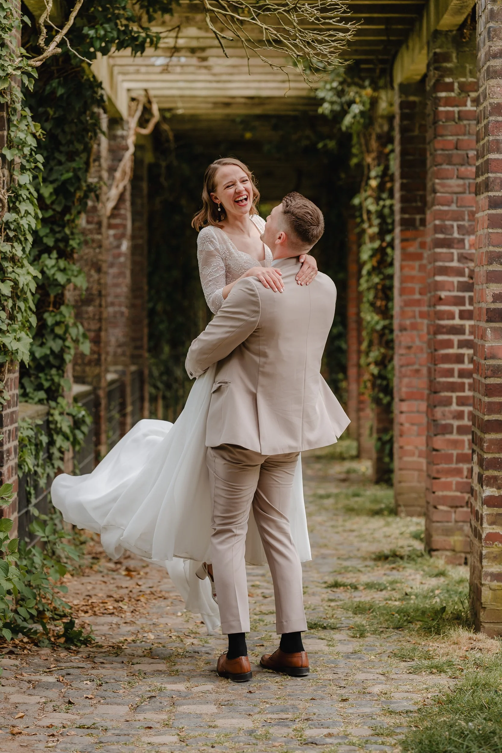 Ein brautpaar bei einer Hochzeit, der Bräutigam hält die Braut in den Armen, sie lachen und wirken glücklich, im Hintergrund ein bewachsener Gartenweg mit Ziegelmauern und einer Pergola.