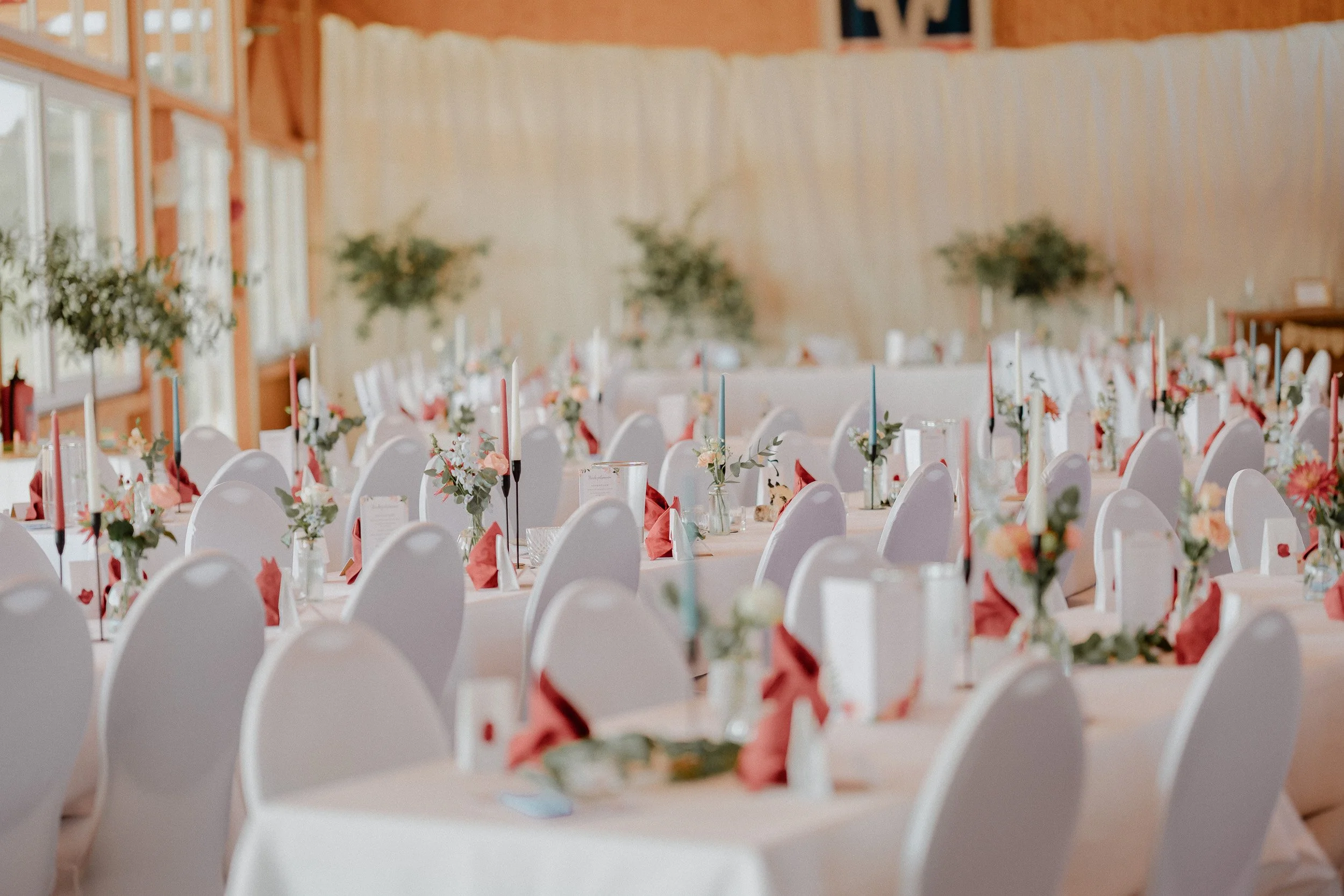 Festlich gedeckter Veranstaltungsraum mit weißen Tischen, Blumenarrangements, Kerzen und Stühlen, vorbereitet für eine Feier.