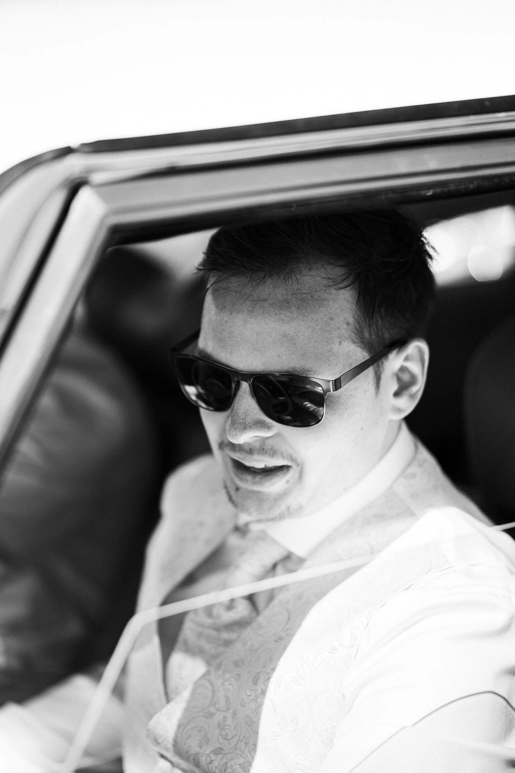 Schwarzer und weißer Foto eines Mannes in Sonnenbrille, sitzend in einem Auto, trägt Anzug und Krawatte.