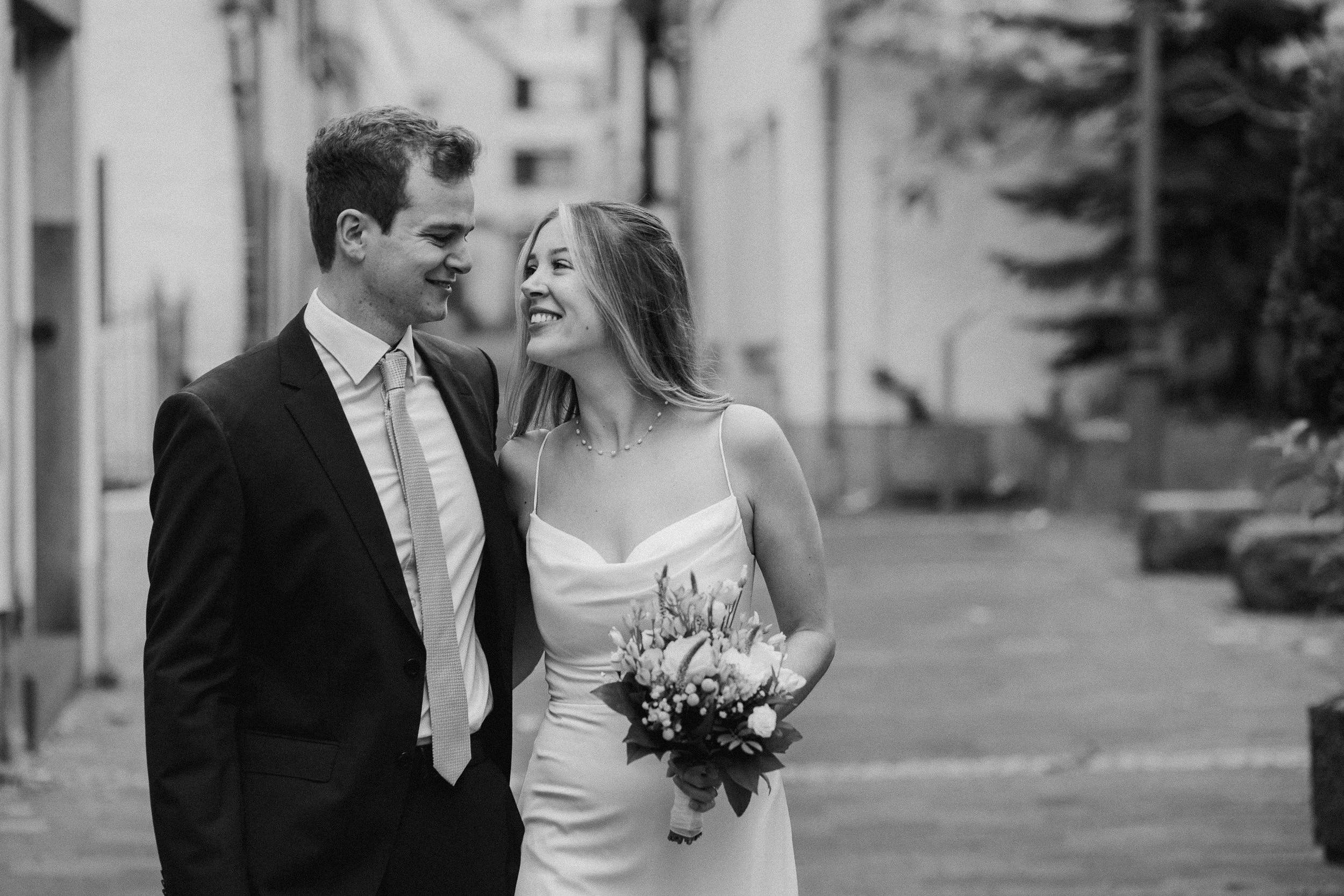 Ein lachendes Brautpaar, ein Mann in Anzug und eine Frau in einem weißen Brautkleid, gehen arm in arm auf einer Straße