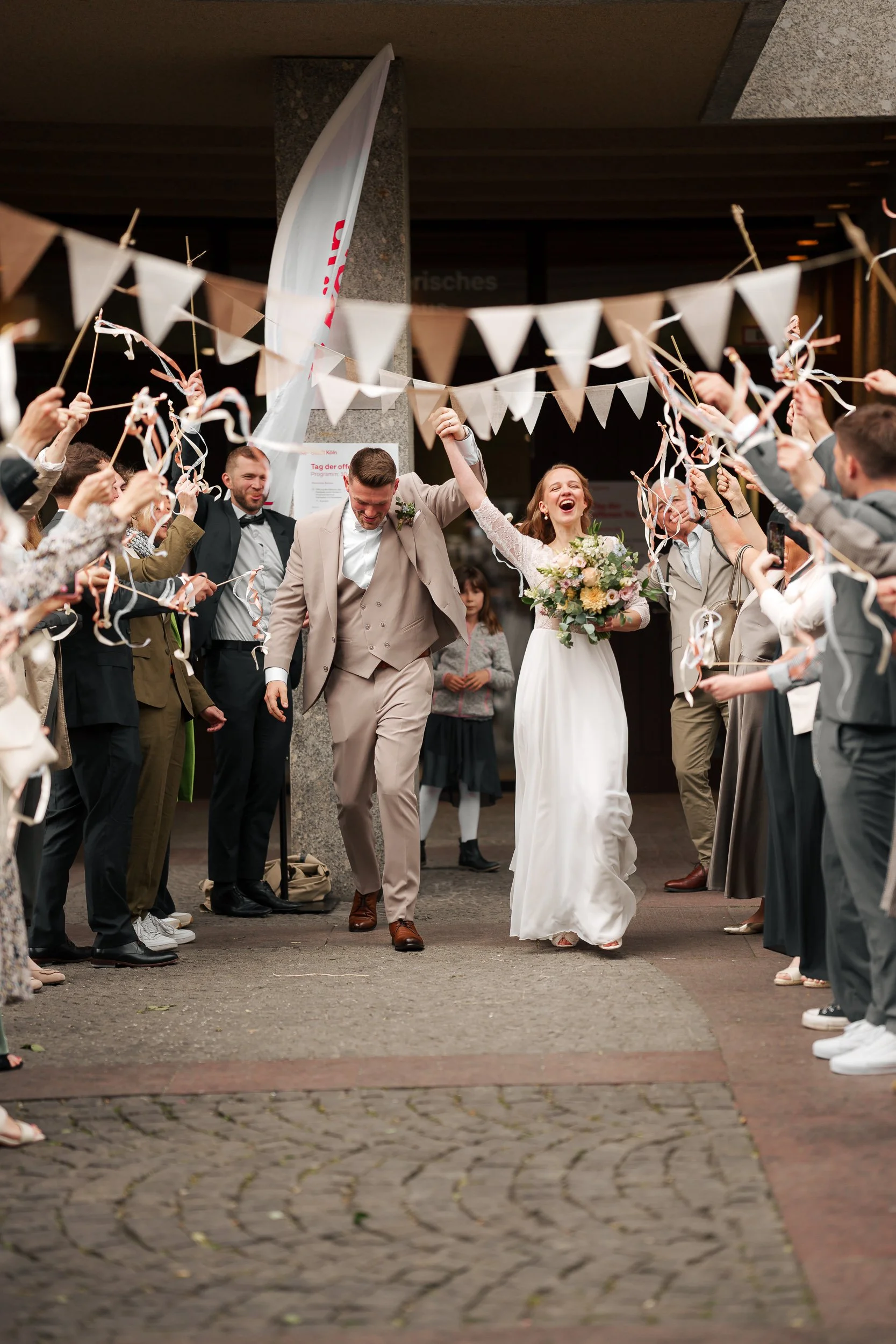 Ein heiratendes Paar läuft lachend durch einen Tunnel aus Gästen, die mit Wimpeln und Papierstreifen winken.