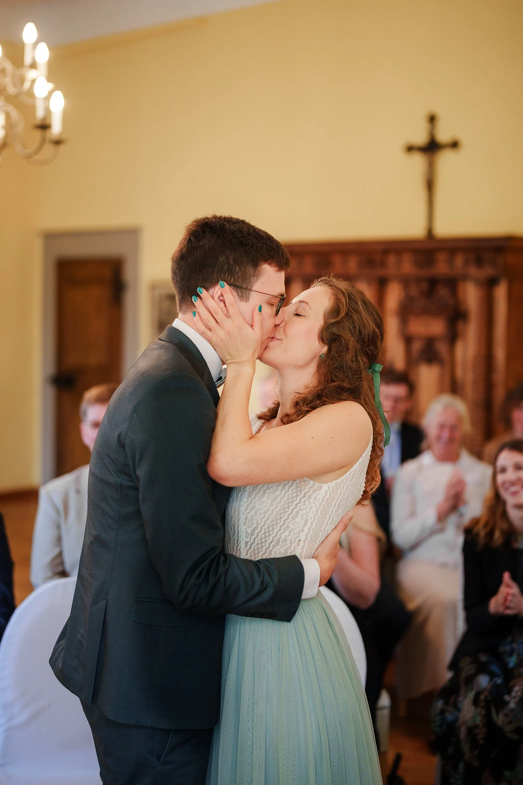 Ein junges Paar bei einer Hochzeitszeremonie küsst sich in einer Kirche, umgeben von anwesenden Gästen.