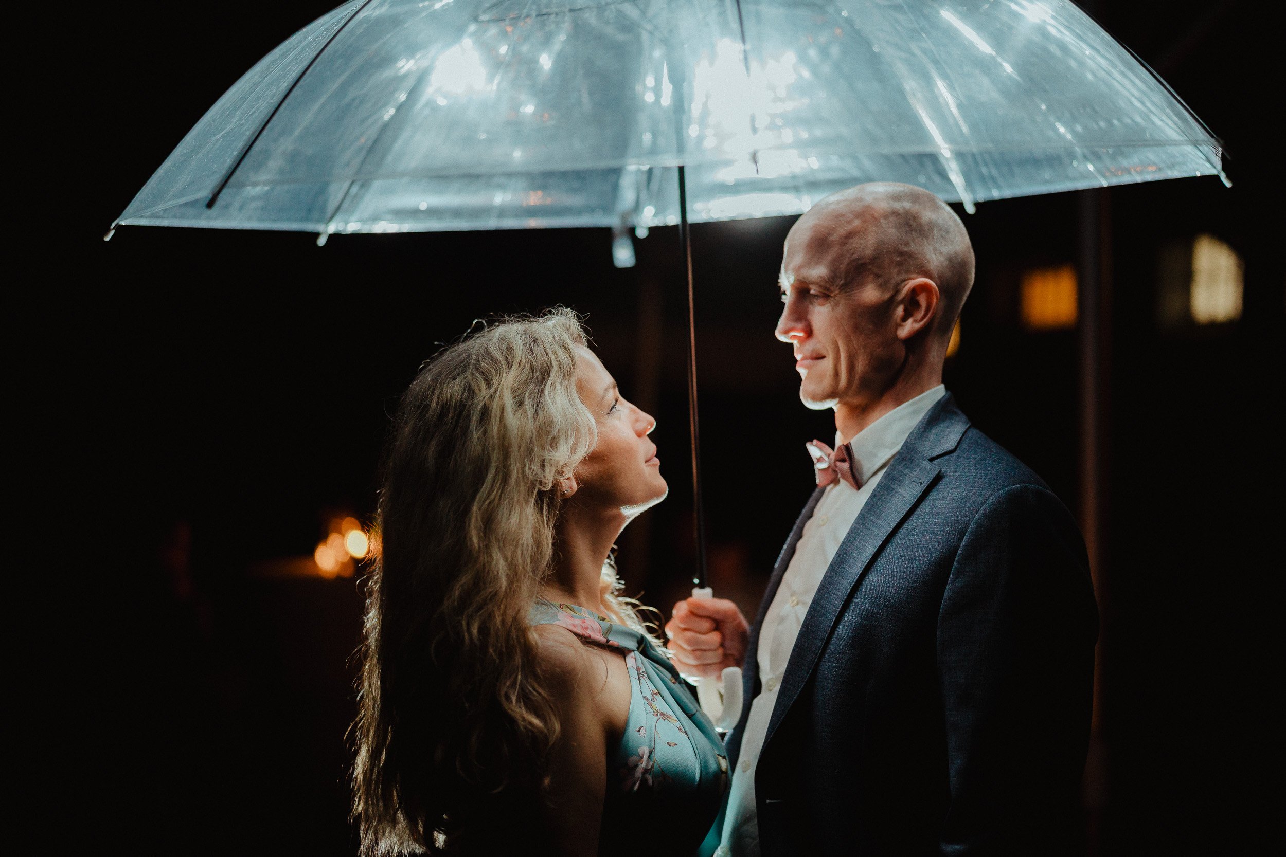 Ein Mann und eine Frau stehen sich bei Nacht unter einem transparenten Regenschirm gegenüber, die beiden schauen sich an, während es regnet.