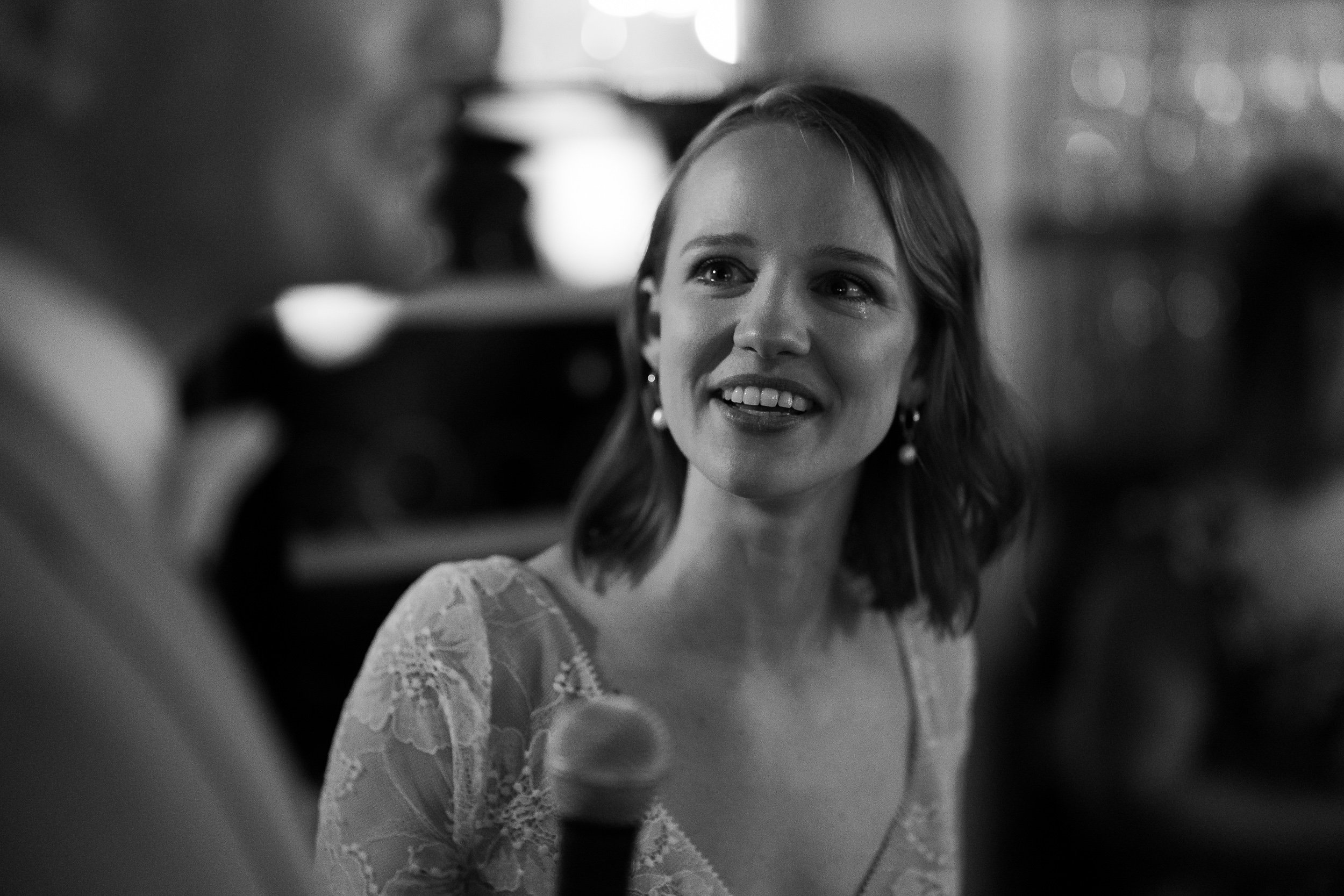 Eine junge Frau mit lockigem, schulterlangem Haar, die lächelnd einem Mann gegenübersitzt, während sie spricht. Im Hintergrund sind unscharfe Lichter, anscheinend bei einer Veranstaltung oder Feier.