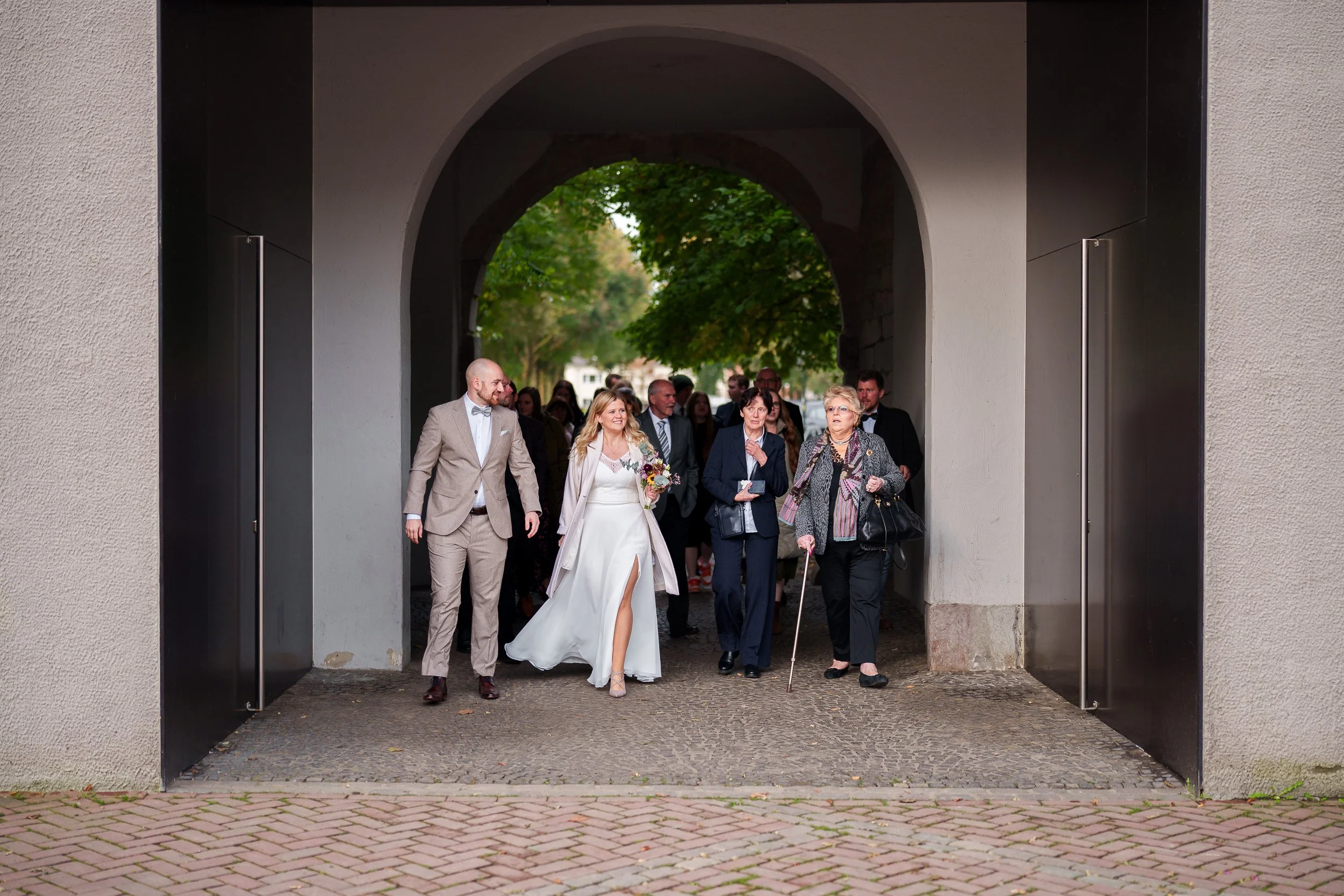 Hochzeitspaar, das durch ein Tor geht, umgeben von Gästen, Frauen und Männer in formeller Kleidung, einige mit Blumen, in einer grünen, urbanen Umgebung.