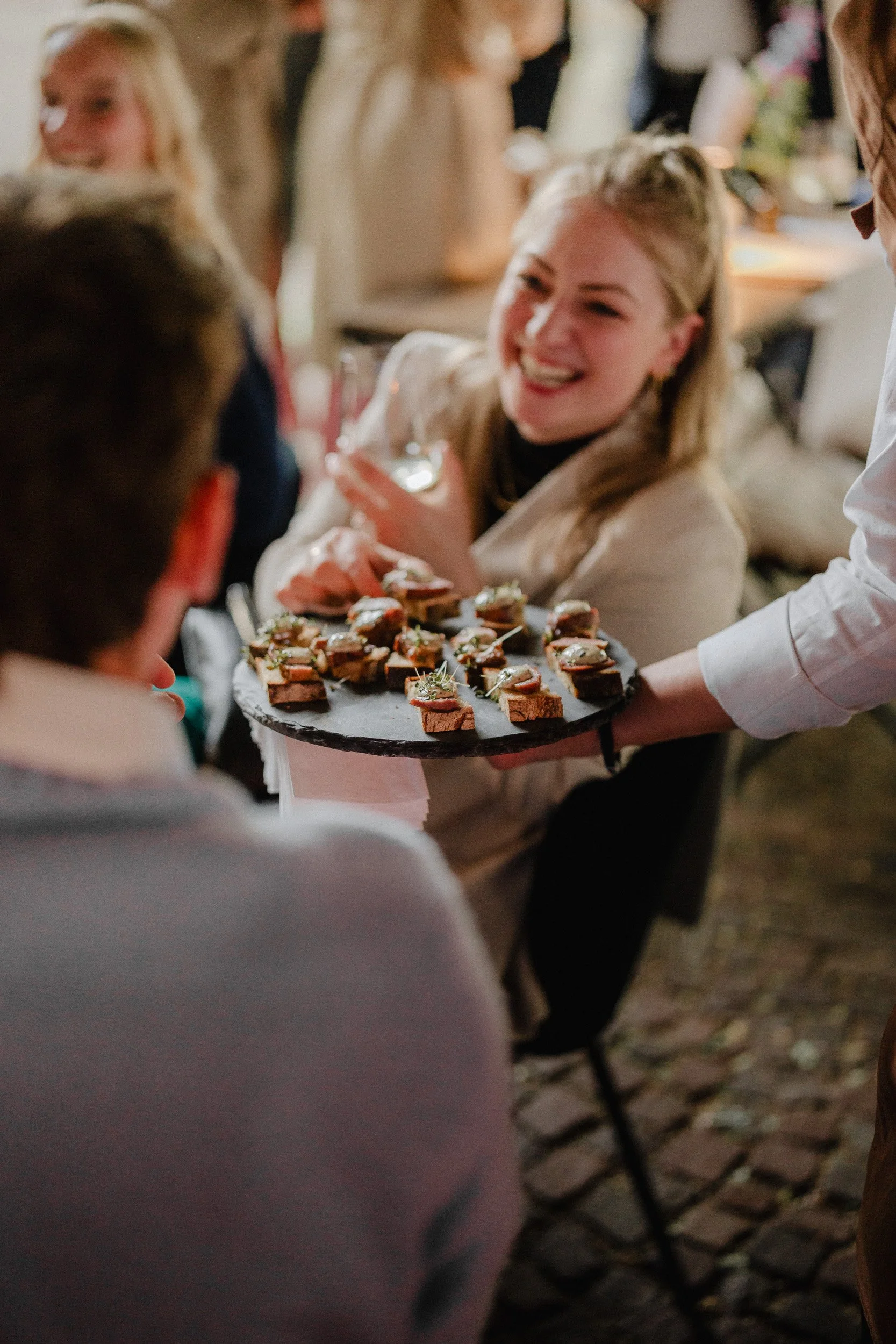 Menschen bei einer Feier, eine Frau bekommt eine Platte mit kleinen belegten Broten serviert, lachende Gäste im Hintergrund.