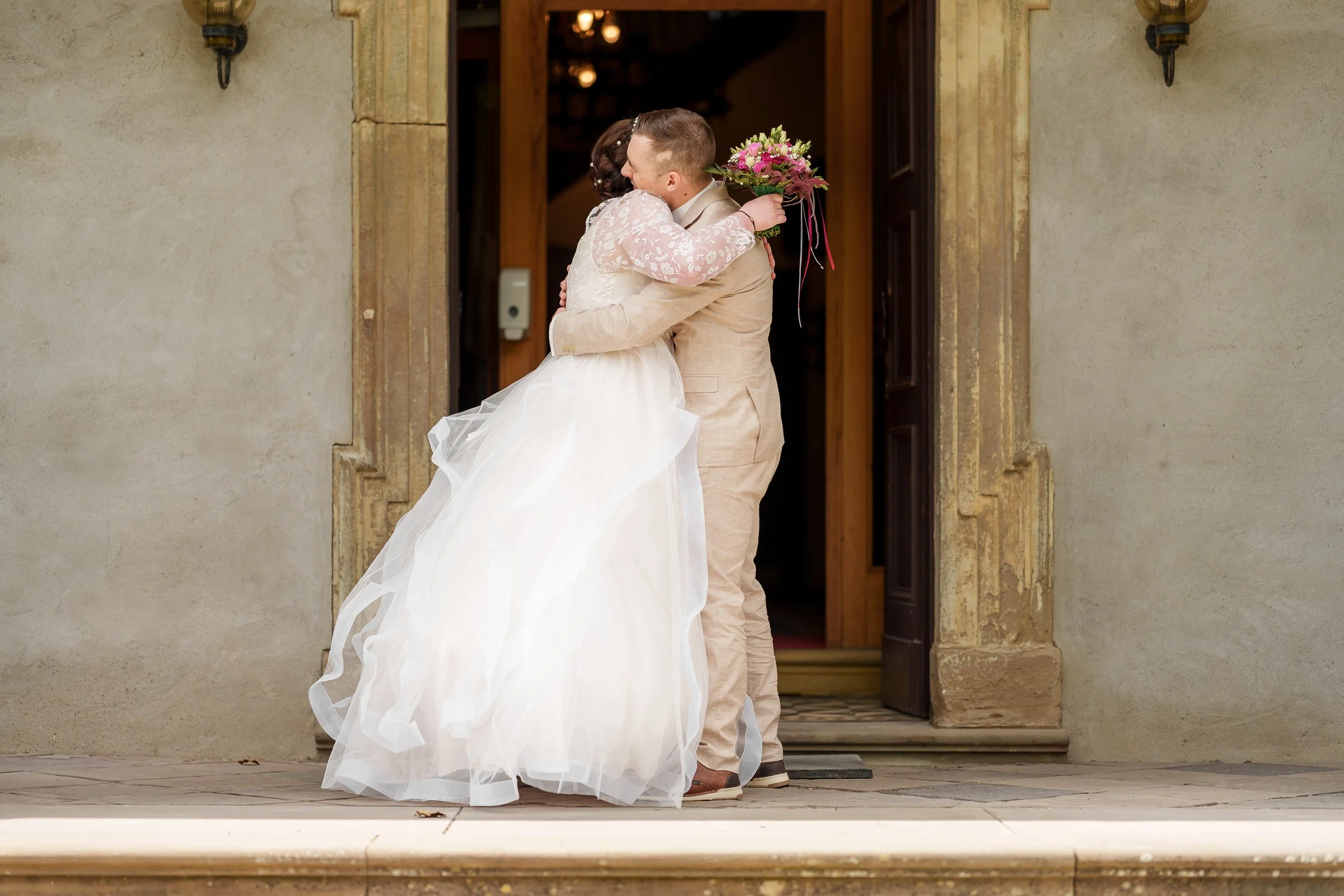 Ein Brautpaar begrüßt sich vor einer Holztür bei einer Hochzeit, die Braut trägt ein weißes Kleid mit Tüll, der Bräutigam trägt einen beige Anzug und hält einen Blumenstrauß.