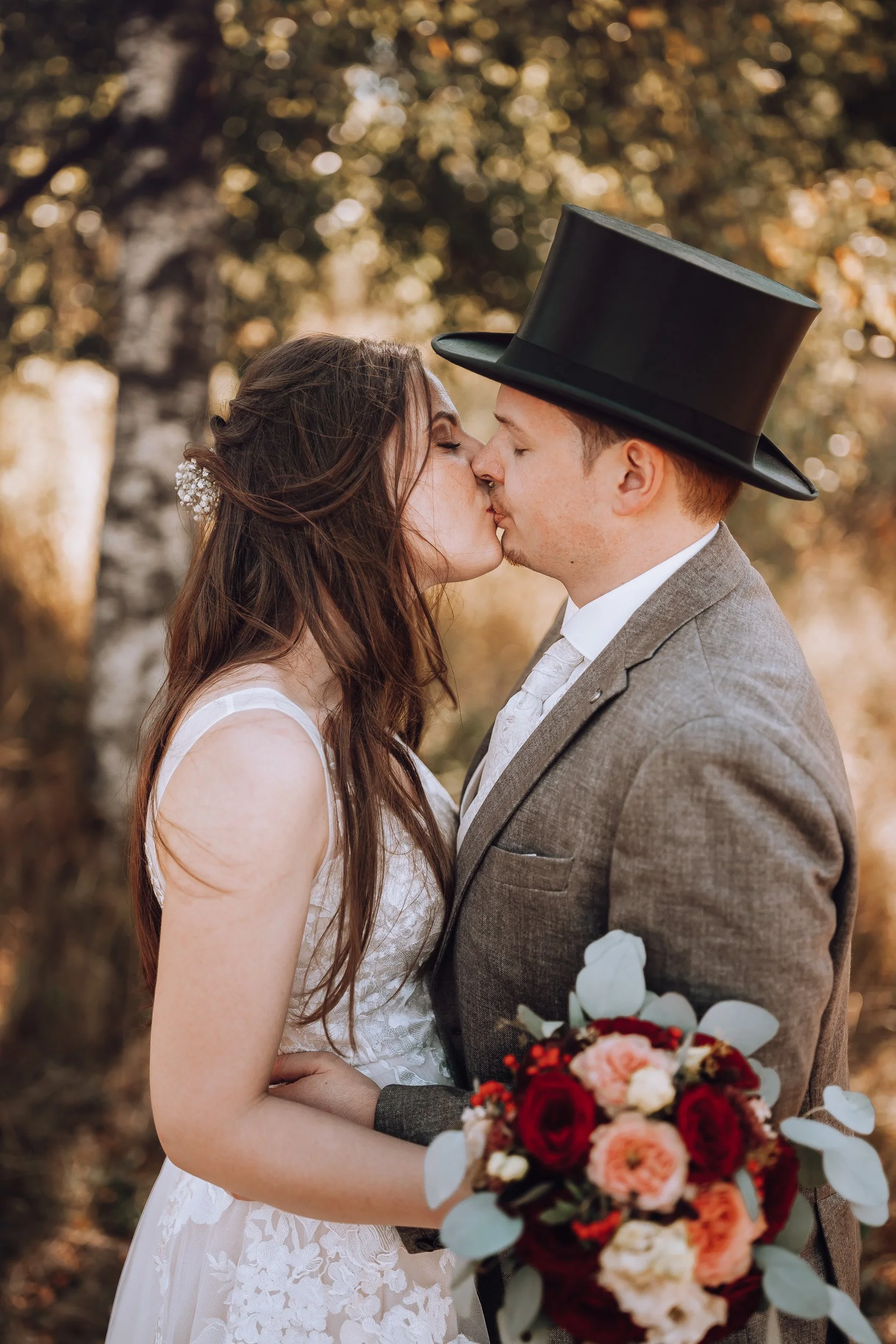 Ein Brautpaar küsst sich im Freien, die Frau trägt ein weißes Hochzeitskleid, der Mann einen braunen Anzug mit einem schwarzen Zylinder. Im Vordergrund hält sie einen Blumenstrauß aus roten und pinken Blumen. Im Hintergrund sind Bäume mit herbstliche