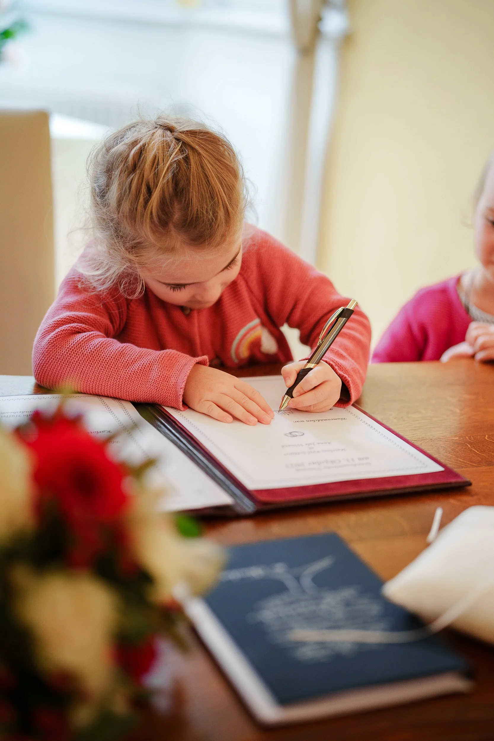 Ein kleines Mädchen schreibt mit einem Stift in einem Foliobuch auf einem Holztisch. Es trägt ein rotes Oberteil und hat blonde, lockige Haare. Im Vordergrund ist eine Blume im Fokus, im Hintergrund eine andere Person.
