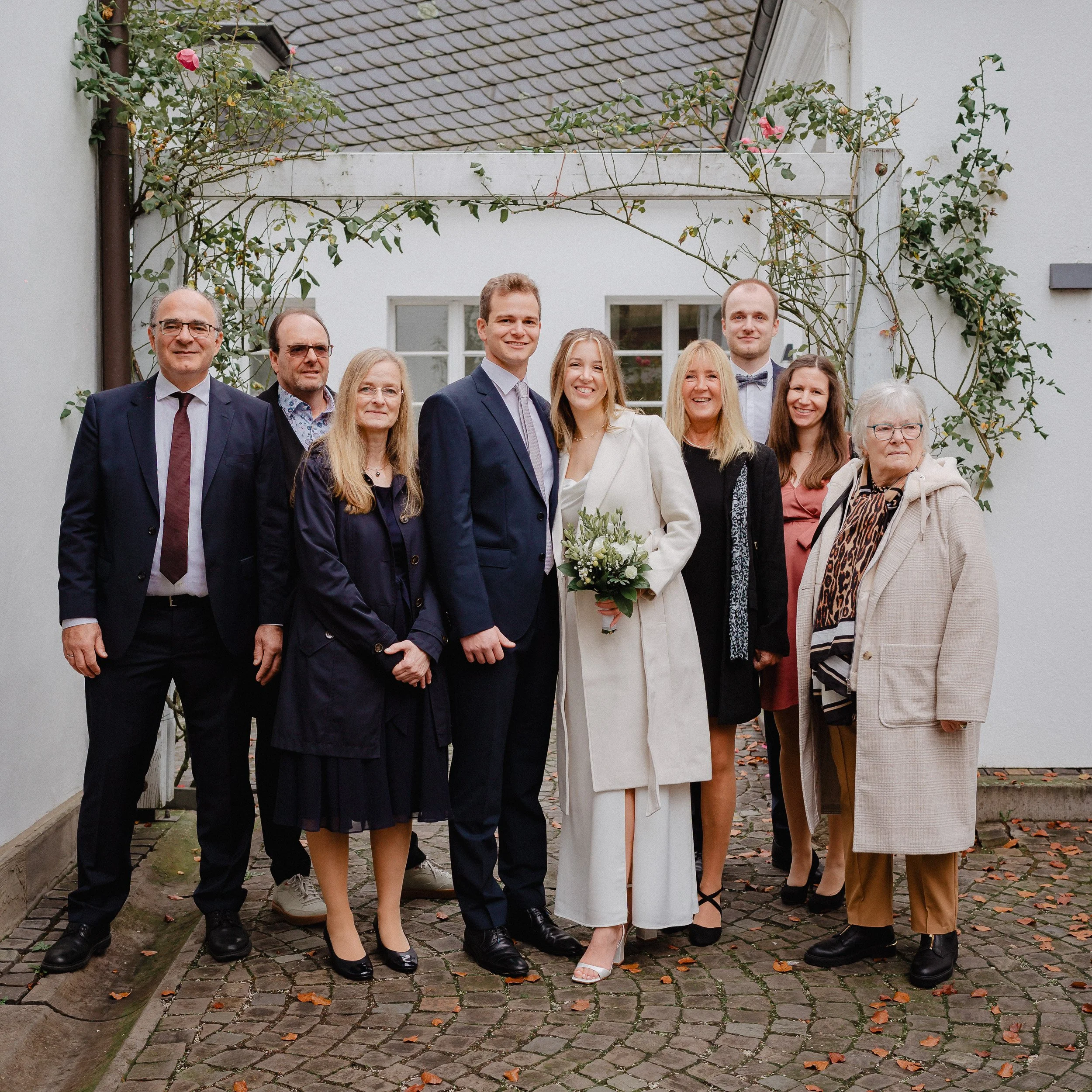 Gruppe von acht Menschen, die bei einer Hochzeitsfeier in einem Hof mit Pflanzranken im Hintergrund posieren.
