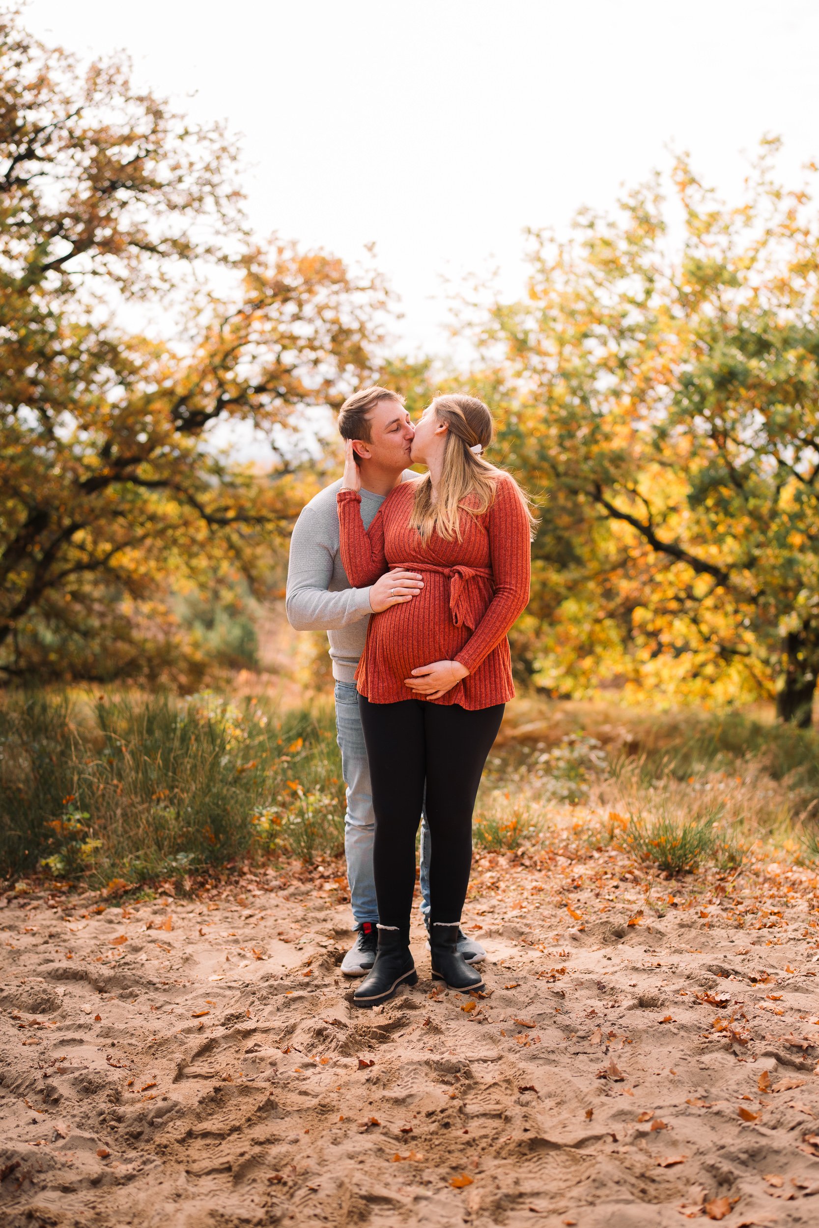 Ein Paar küsst sich im Herbstpark, die Frau ist schwanger, die Bäume im Hintergrund haben orangefarbene und gelbe Blätter.