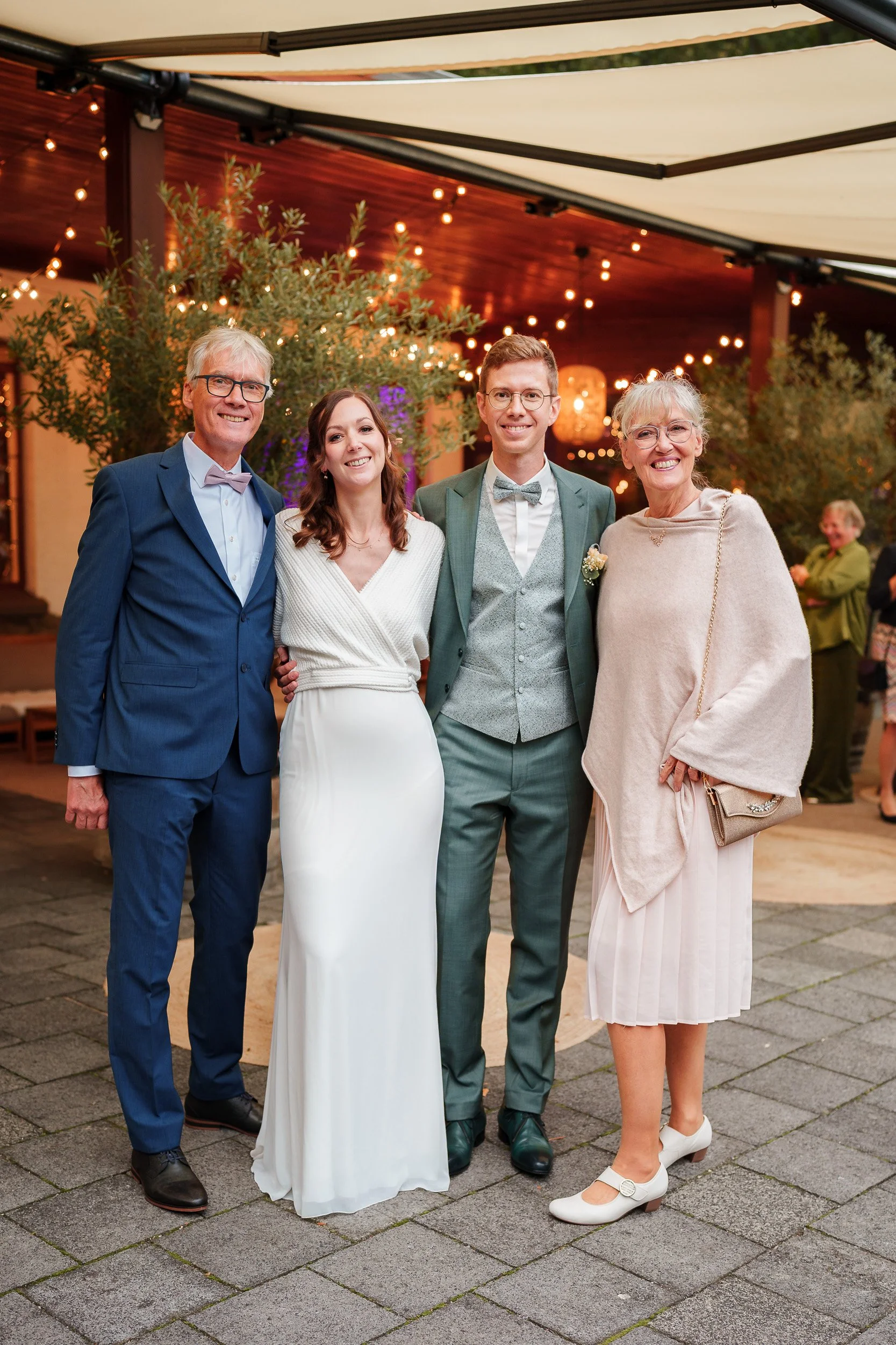 Fünf Menschen in formeller Kleidung bei einer Feier, darunter ein Brautpaar und zwei ältere Personen, im festlich dekorierten Innenraum.