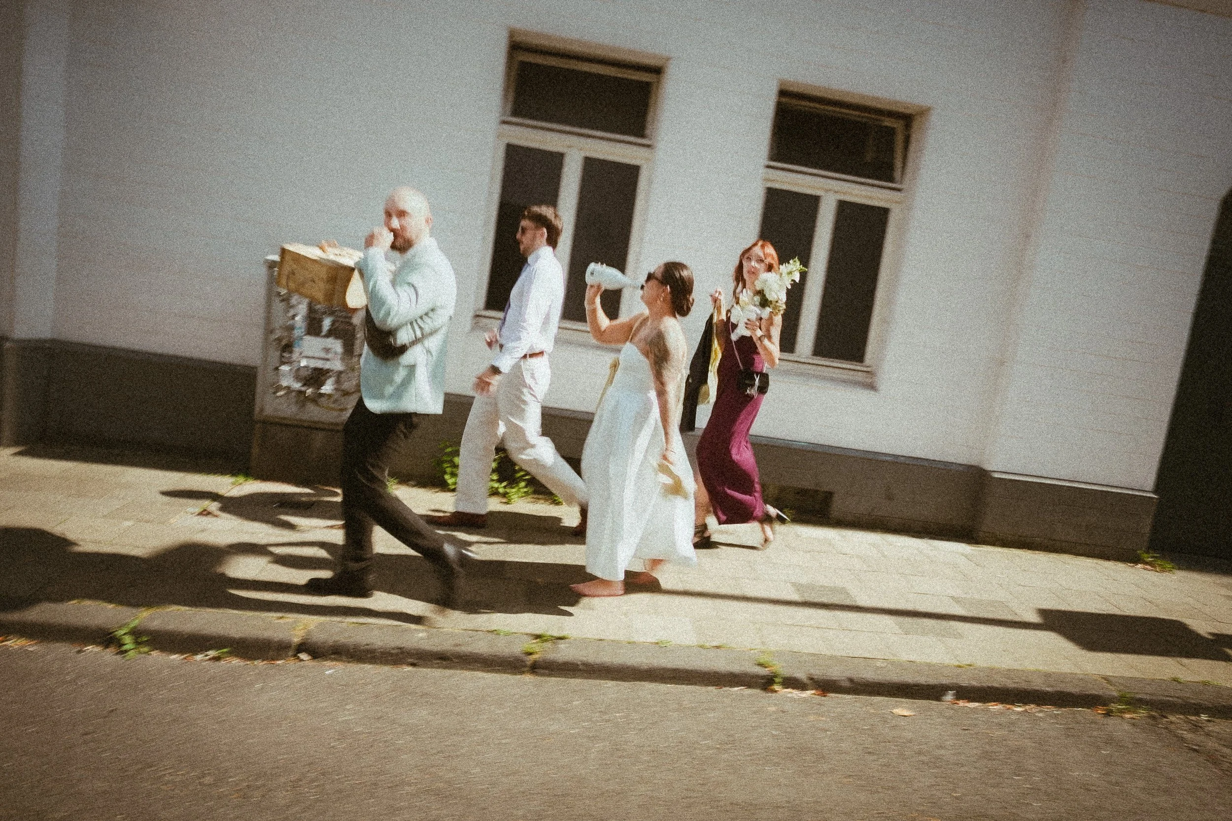 Fünf Menschen gehen an einem Gebäude auf dem Bürgersteig vorbei. Eine Frau trägt ein weißes Kleid und hat Tattoos, hält eine Wasserflasche, eine andere Frau trägt einen dunklen Anzug und hält einen Blumenstrauß, ein Mann trägt einen weißen Anzug, ein
