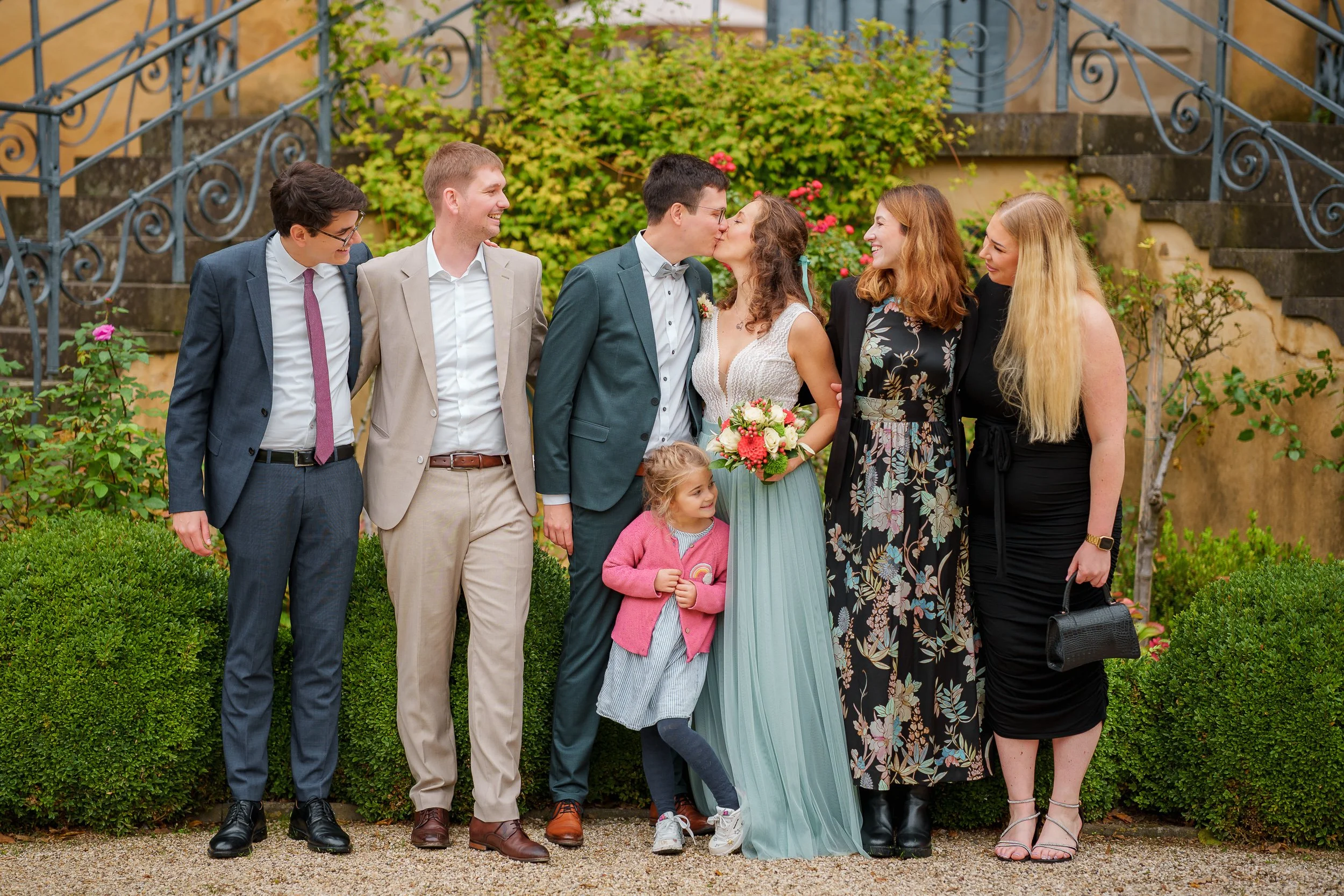 Eine Gruppe von acht Menschen, darunter ein Paar, das sich küssend in der Mitte, bei einer Hochzeit im Freien, umgeben von einem grünen Garten mit Blumen, Fotos machen.