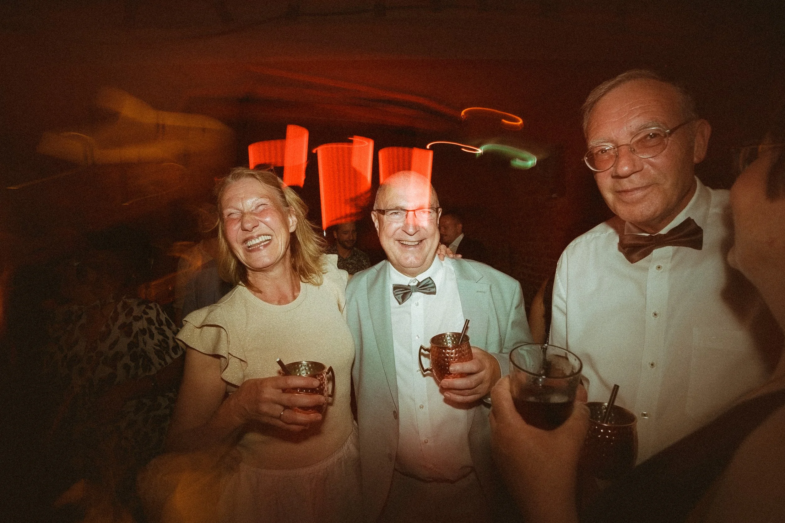 Drei Menschen auf einer Party, lachen und halten Getränke in der Hand, im Hintergrund sind bunte Lichter und weitere Gäste.
