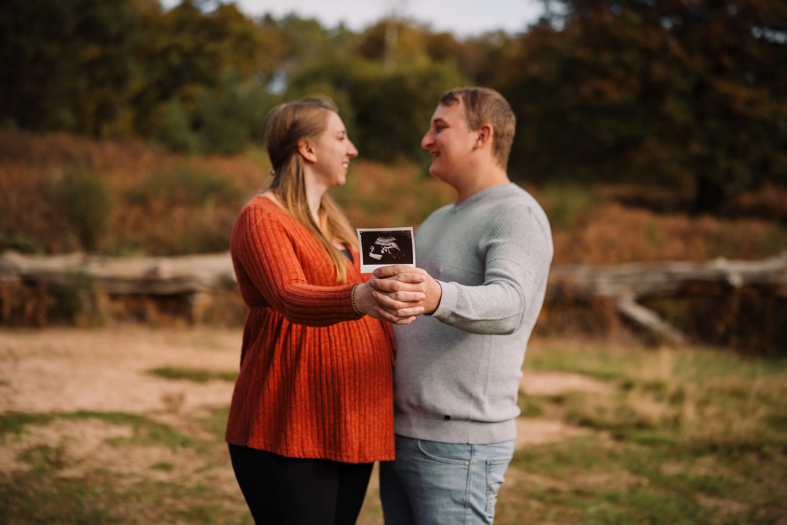 Ein Paar hält ein Ultraschallbild im Freien, umgeben von Herbstlaub und Bäumen.